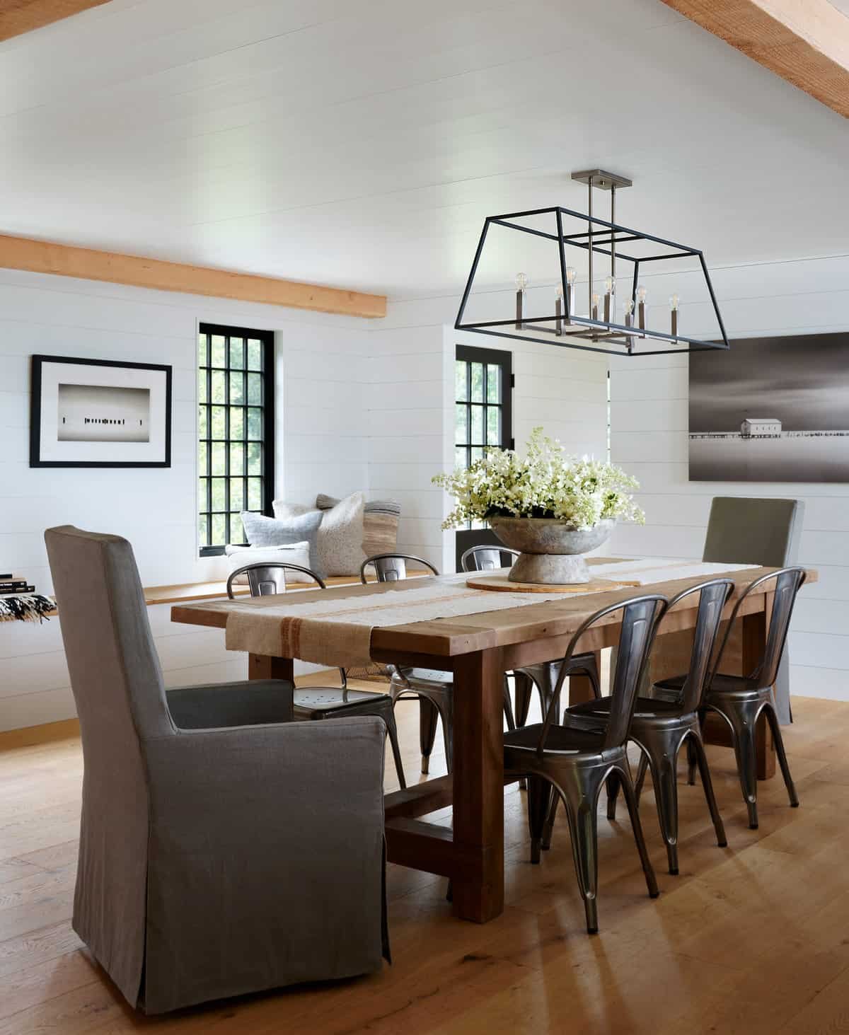 Dining room with a wooden table, six metal chairs, a gray upholstered armchair, a flower arrangement, and wall art, with two windows and a modern chandelier.