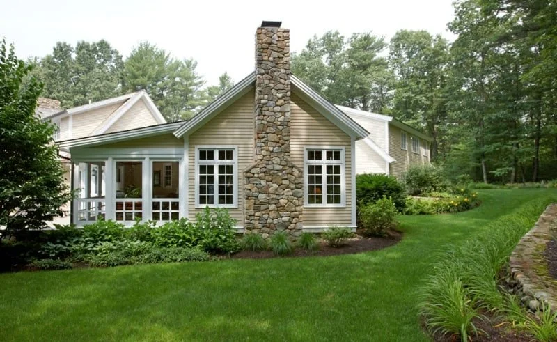 A house with a stone chimney, surrounded by green lawns and bushes, in a wooded area.