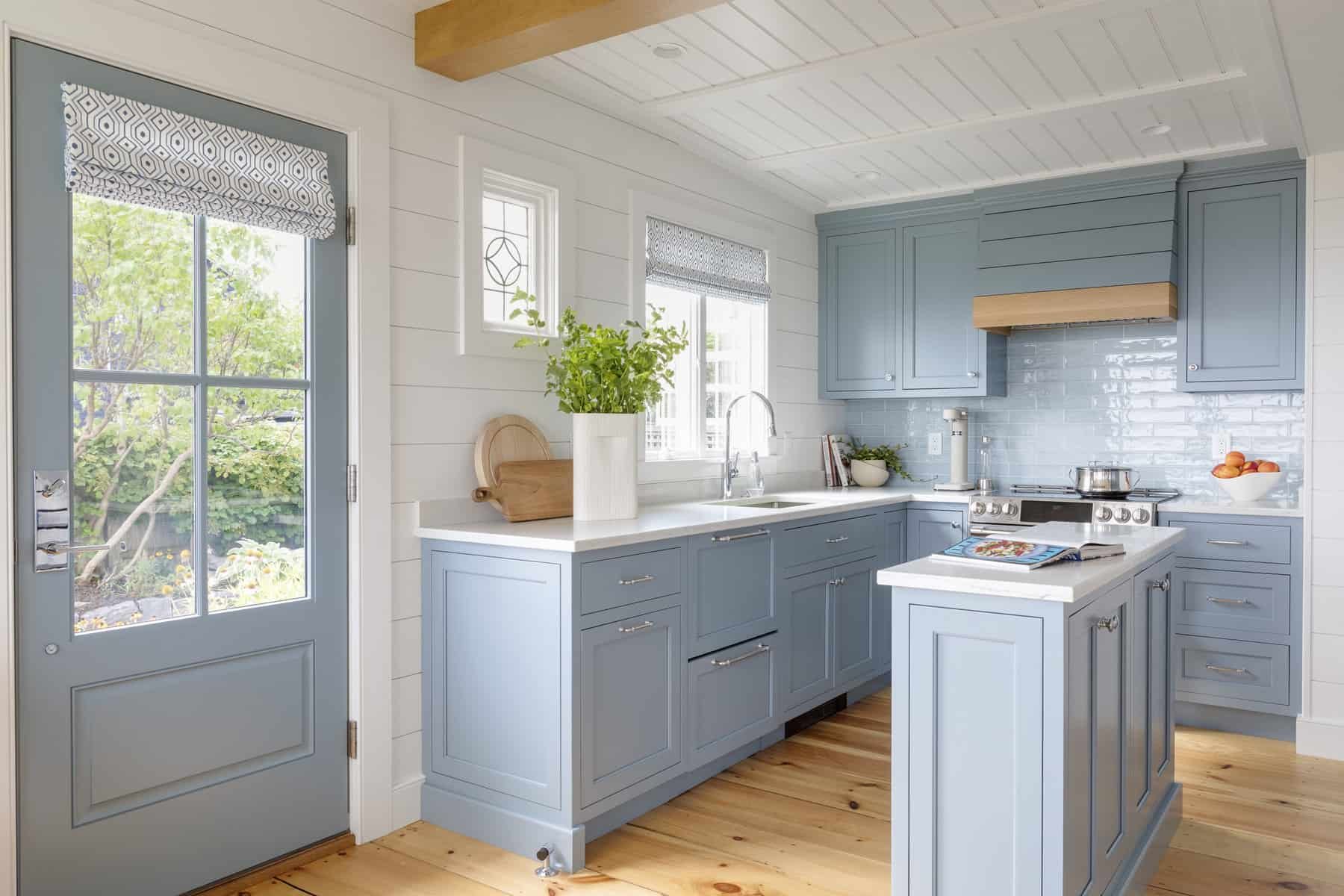 A kitchen with blue cabinets, white countertops, a door with glass panes leading outside, and windows with patterned blinds; decorated with green plants, a cookbook, and kitchen utensils.