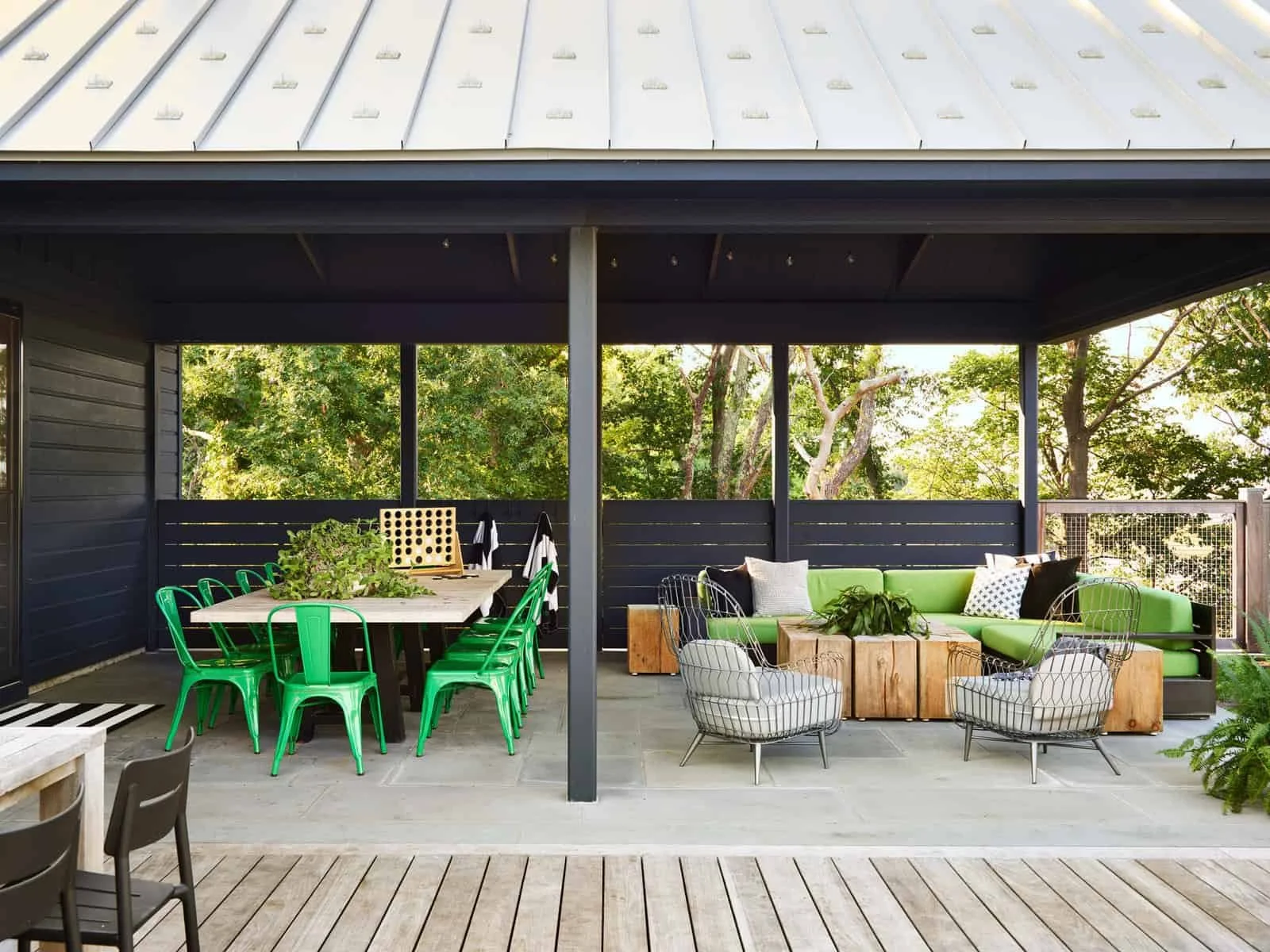 An outdoor patio area with black railing, green chairs around a wooden table, a green sofa with pillows, and small outdoor tables, surrounded by trees.