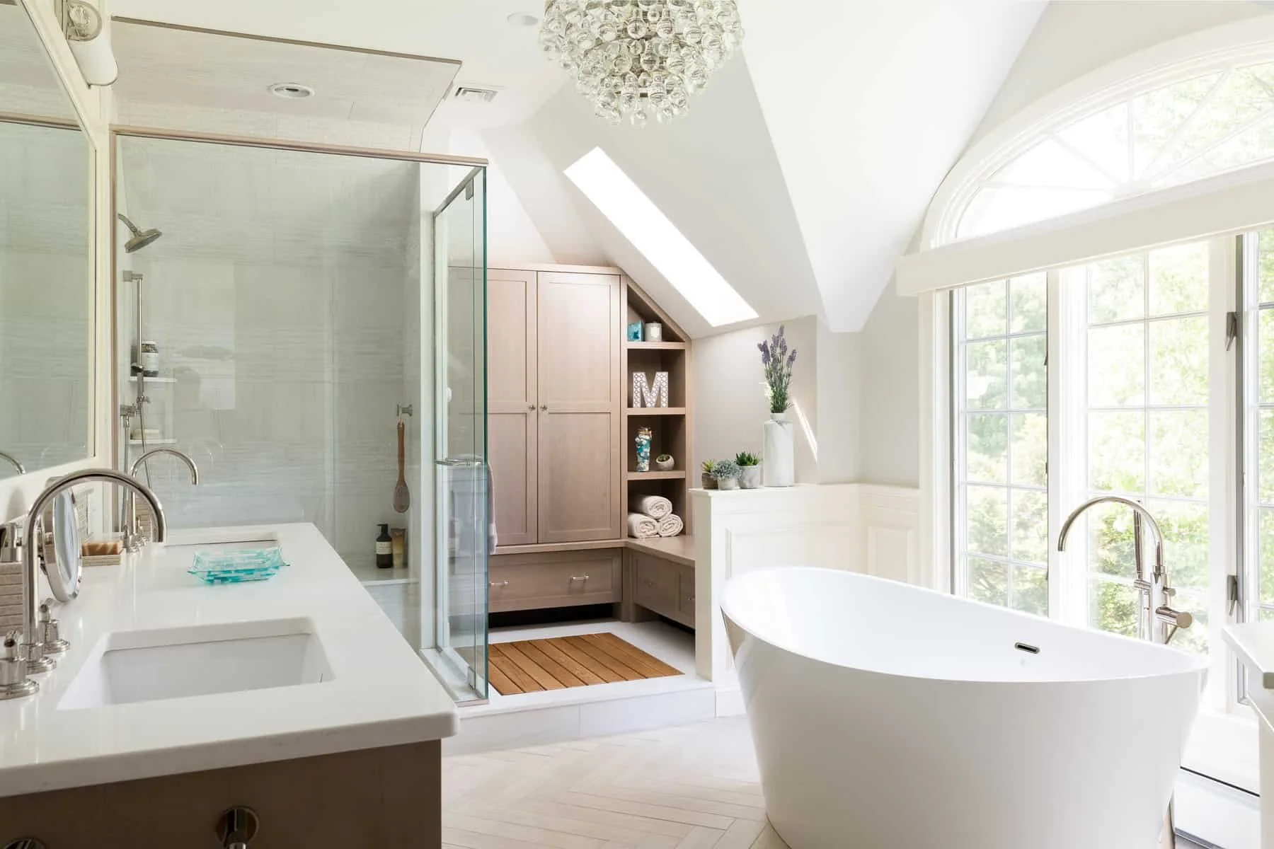 Bright bathroom with large arched window, free-standing bathtub, glass-enclosed shower, double vanity with sinks, and wooden storage cabinetry with decorative items and plants.