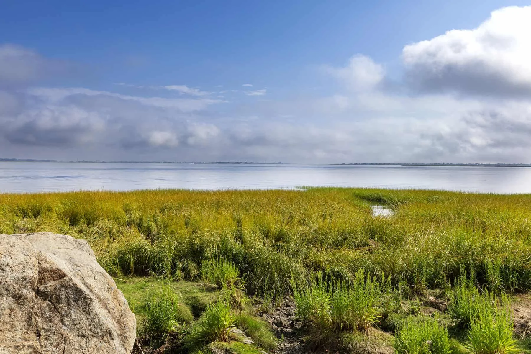 A peaceful landscape showing a grassy marsh with a small waterway, leading to a body of water under a partly cloudy sky.