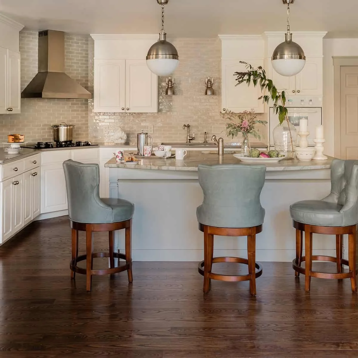 A modern kitchen with white cabinets, marble countertops, and a wooden floor. There are three upholstered bar stools at the counter, with a vase of flowers and decorative candles on the counter. Stainless steel appliances like a stove, range hood, an