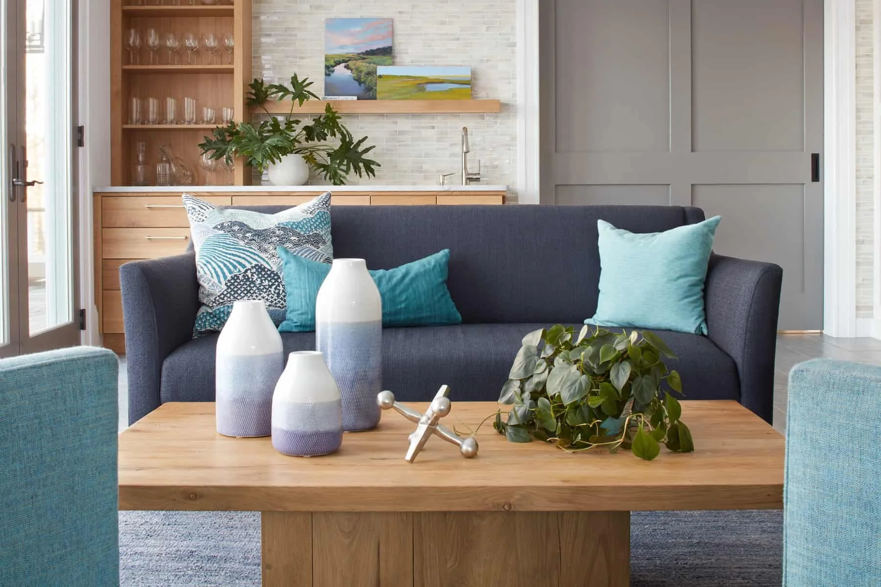 Living room with a dark blue sofa, light blue and patterned throw pillows, a wooden coffee table with decorative vases and greenery, and a background featuring a kitchenette with artwork, a plant, and a sliding door.