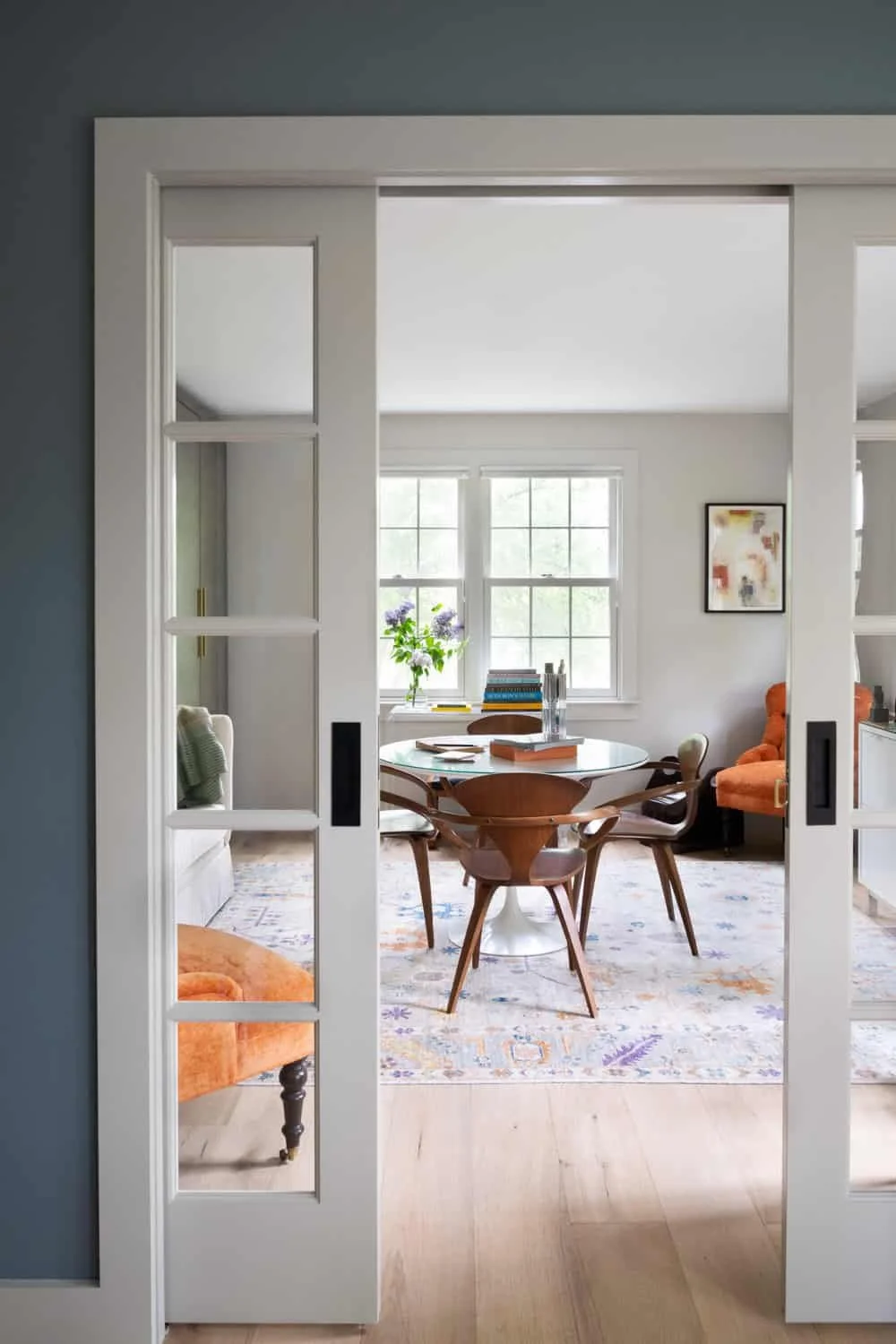 View of a cozy dining room seen through sliding glass door with multiple window pane sections; contains a round table, wooden chairs, and a window with a vase of flowers.
