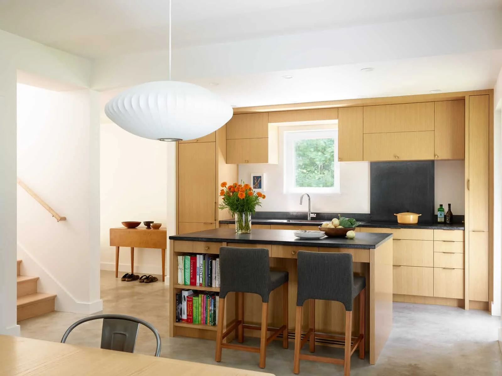 Modern kitchen with wooden cabinets, black countertop, and a small island with books and chairs. A white pendant light hangs from the ceiling, and a window provides natural light. Fresh flowers in a vase are on the island.