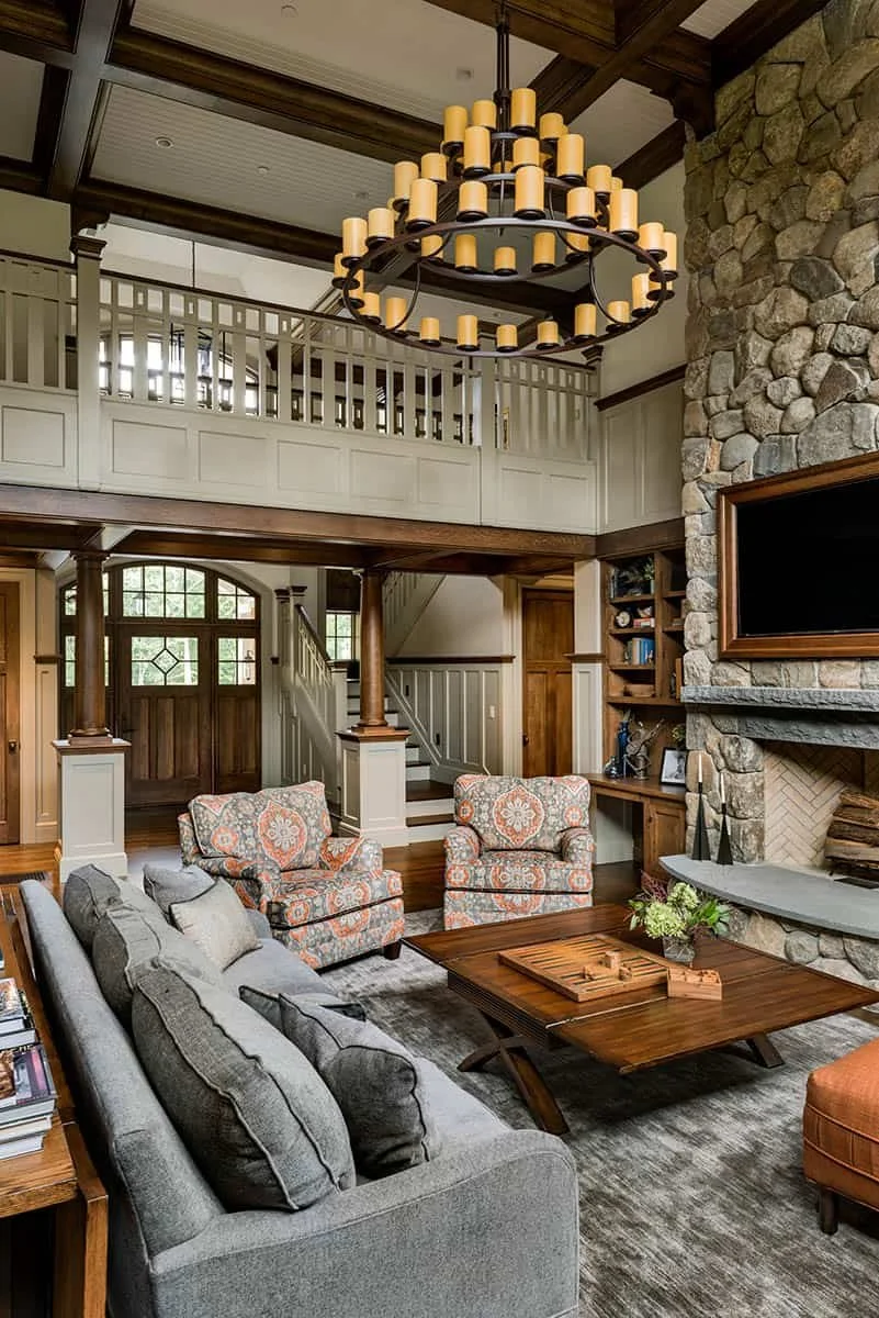 Living room with a high ceiling, stone fireplace, large chandelier, and an upstairs balcony. Contains a gray sofa, two patterned armchairs, a wooden coffee table, and built-in wood shelves.