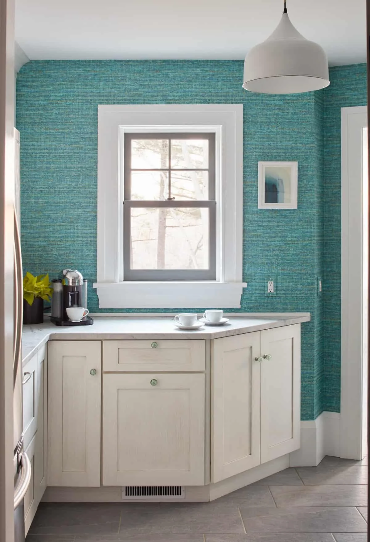 Kitchen with turquoise patterned wallpaper, a window with white trim, white cabinets, a coffee machine, two cups on saucers, and a framed picture on the wall.