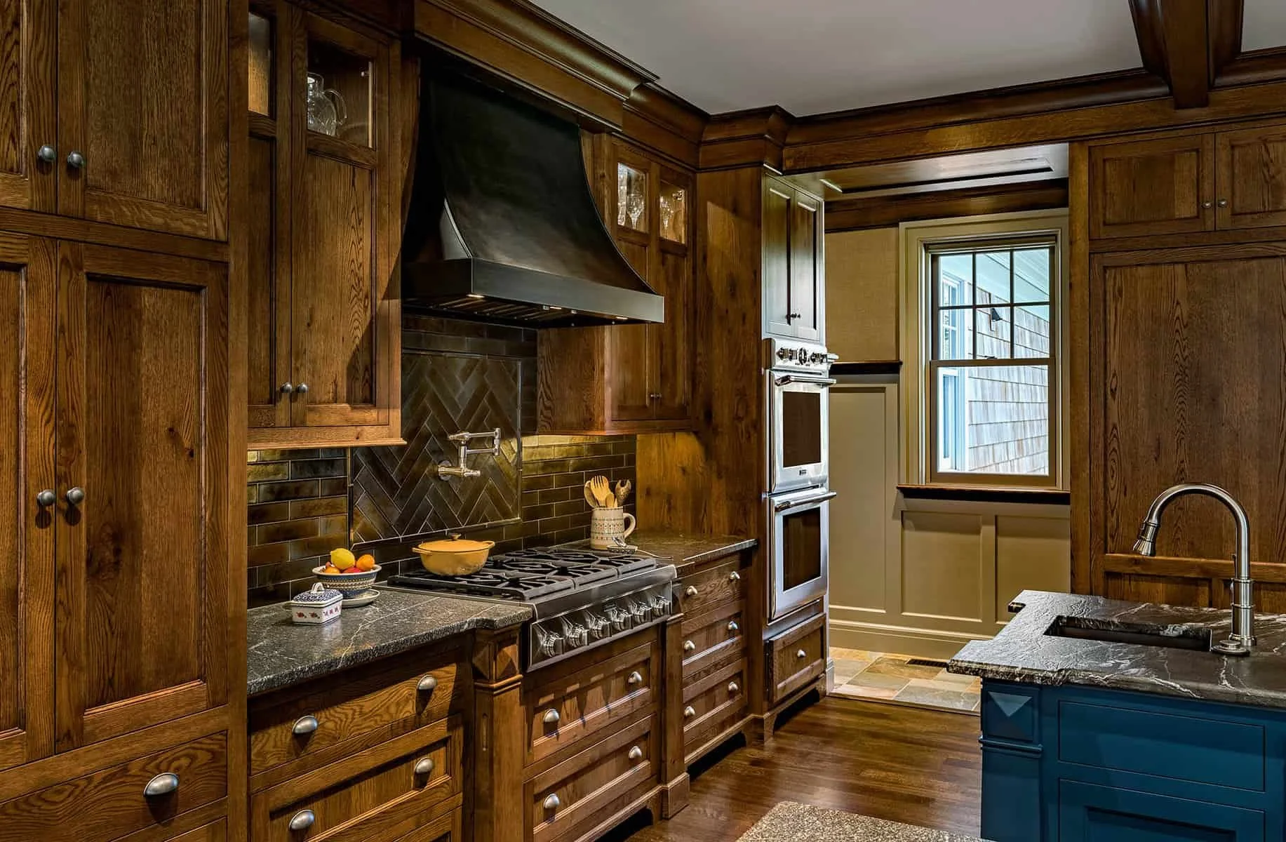 A kitchen with dark wood cabinets, a black range hood, a stove, a window, and a blue kitchen island.
