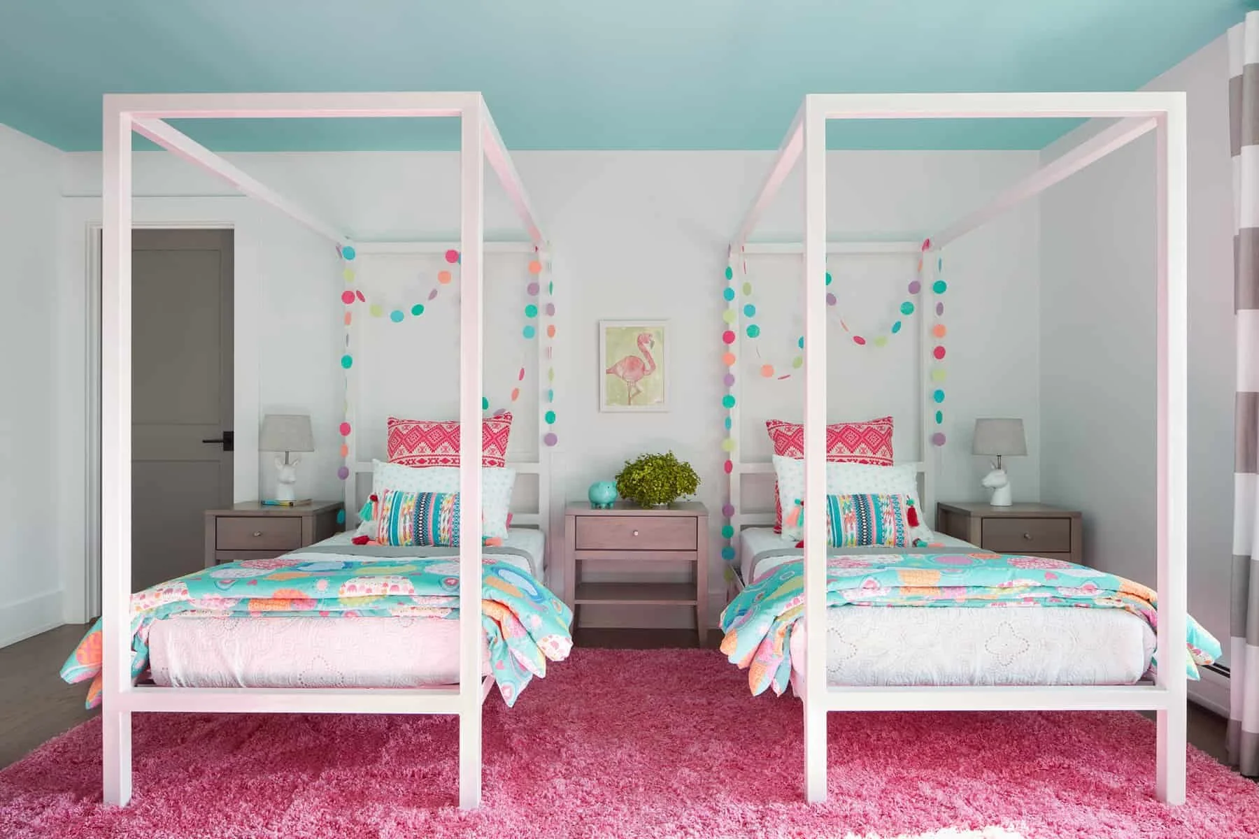 A brightly decorated bedroom featuring two twin beds with colorful bedding and pillows, a pink shaggy rug, white side tables with lamps, and colorful paper garlands hanging from the ceiling. A framed pink flamingo artwork is centered on the wall betw