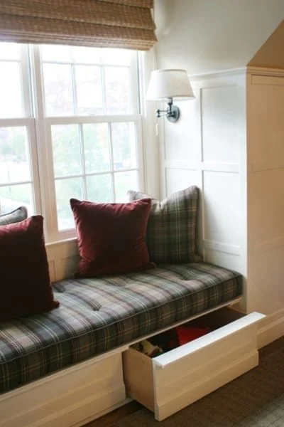 A built-in window seat with plaid cushion, red pillows, and a white drawer underneath near a large window with blinds, a white wall-mounted lamp, and white wall paneling.