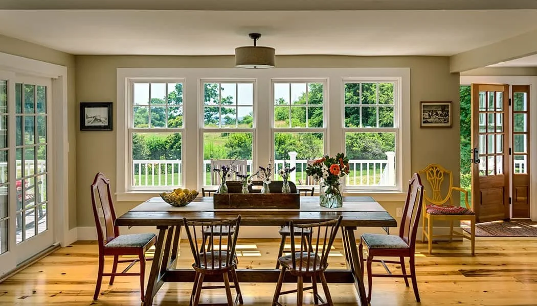 Bright dining room with large windows overlooking a green yard, wooden table with chairs, and flowers in a vase.