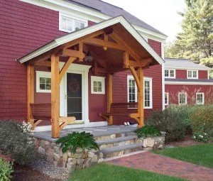 Front porch of a house with a wooden covered entrance, bench, and stone steps, surrounded by greenery.
