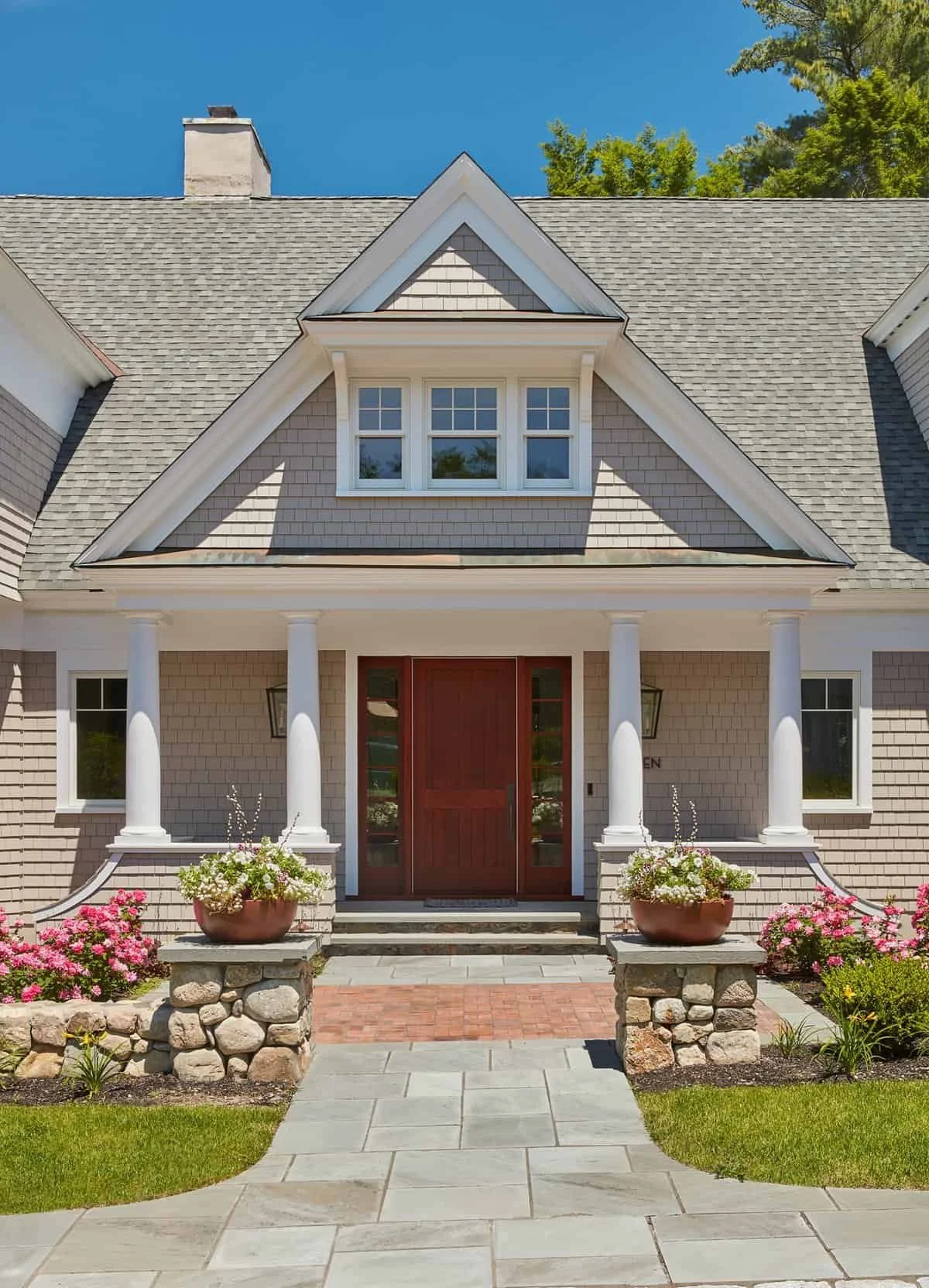 Front entrance of a house with a red door, white columns, and flowerpots with colorful flowers, green grass, and a stone pathway leading to the door.