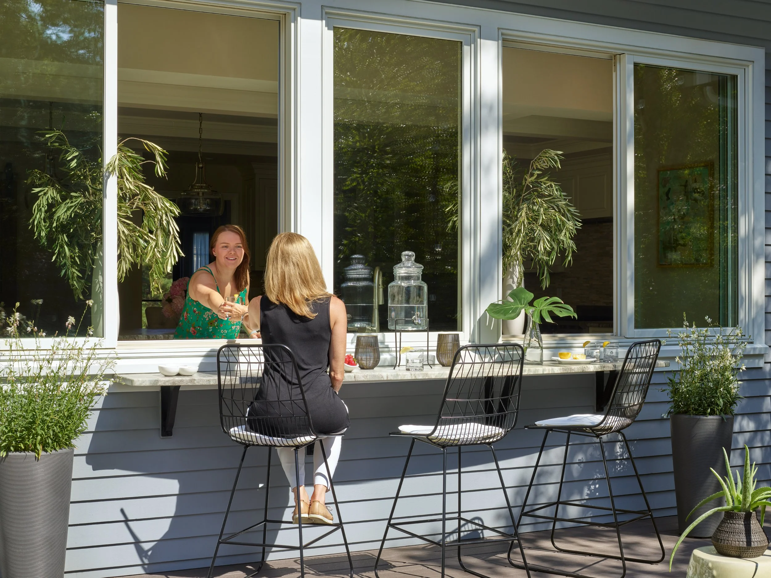 Two women having a conversation at an outdoor dining area, seen through a large glass window with greenery in the background. The woman with red hair is smiling and wearing a green floral dress, while the other with blonde hair is sitting with her ba