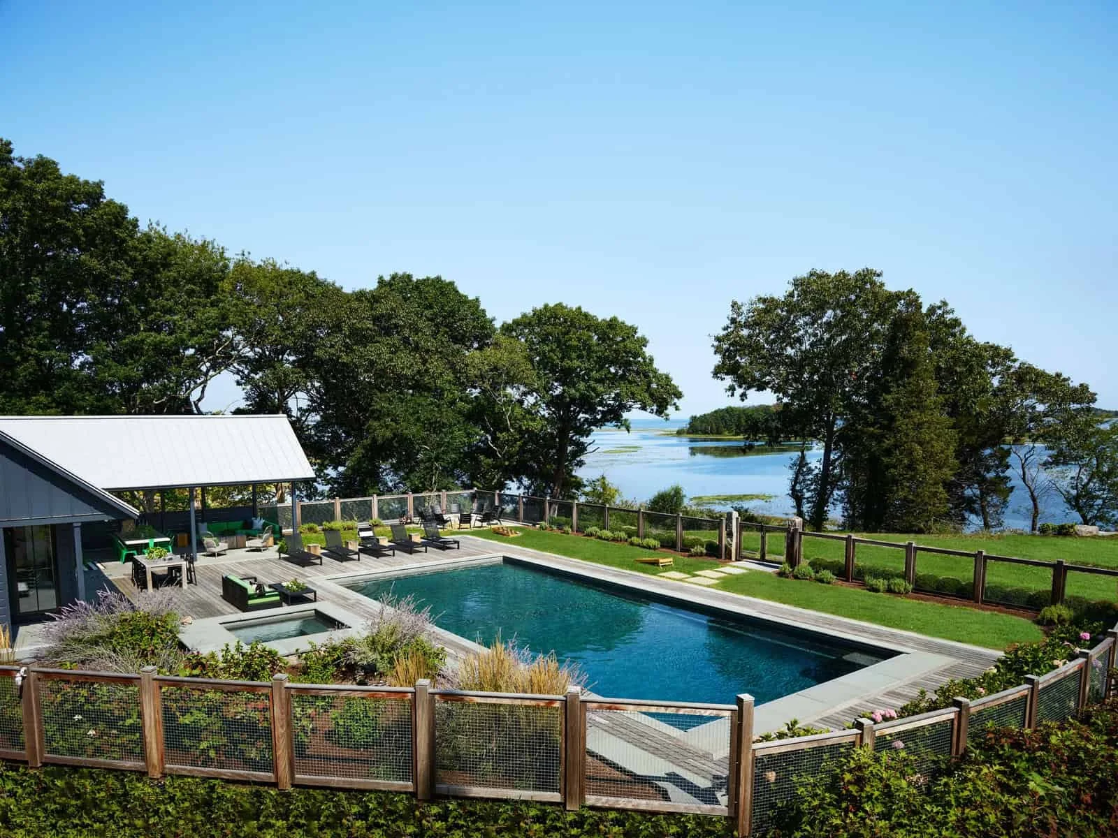 A backyard with a swimming pool, lounge chairs, and a view of a river surrounded by trees and green landscape under a clear blue sky.