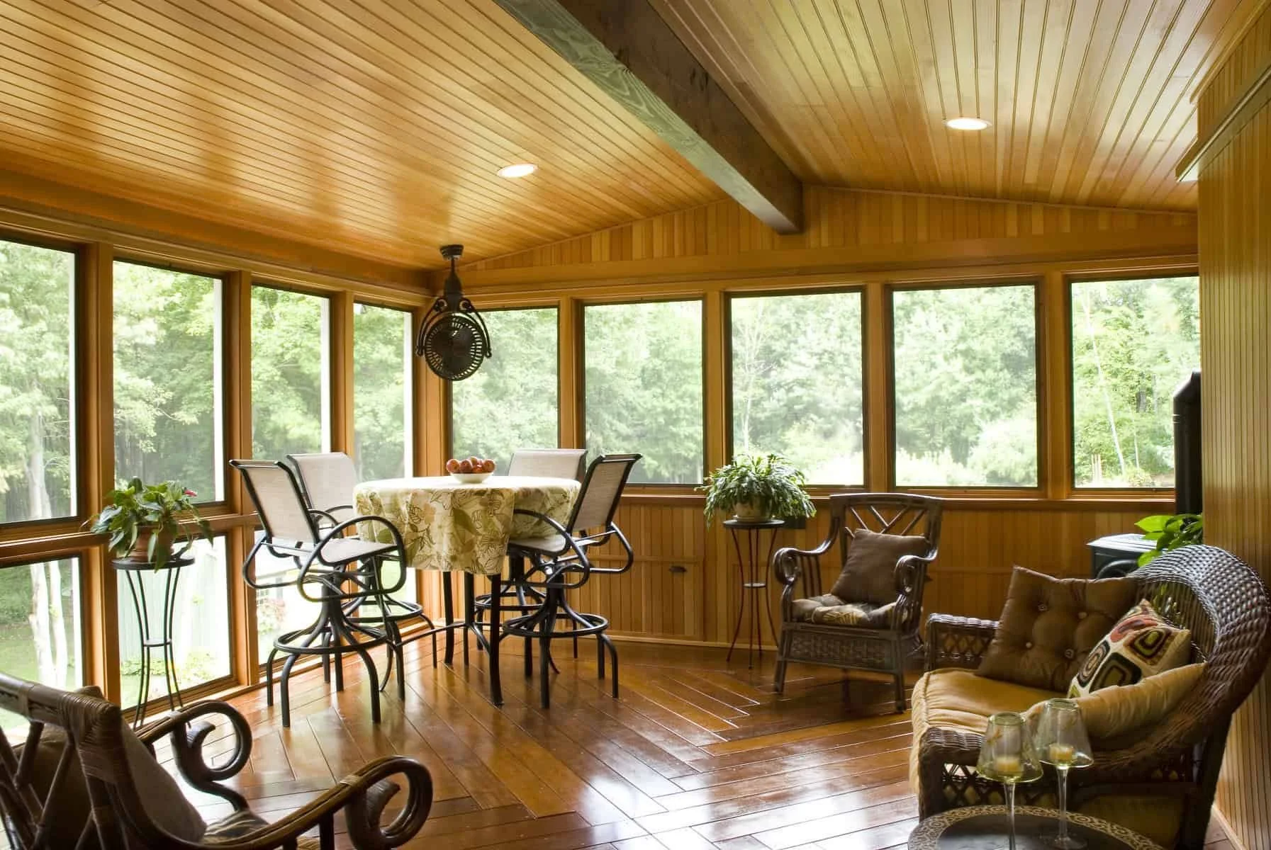 A sunroom with wooden paneled walls and ceiling, large windows showing a green outdoor landscape, a round table with a floral tablecloth, four chairs around the table, and some potted plants inside.