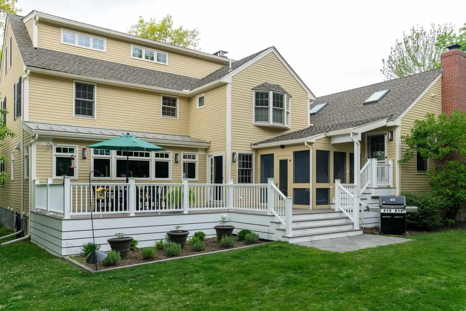 Backyard view of a yellow, multi-story house with a white deck, outdoor furniture, umbrella, potted plants, a grill, and lush green lawn.