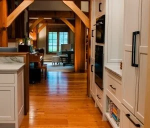 View of a cozy kitchen with white cabinets, hardwood floors, and a wooden beam ceiling, leading to a bright living area with large windows and a table with chairs.