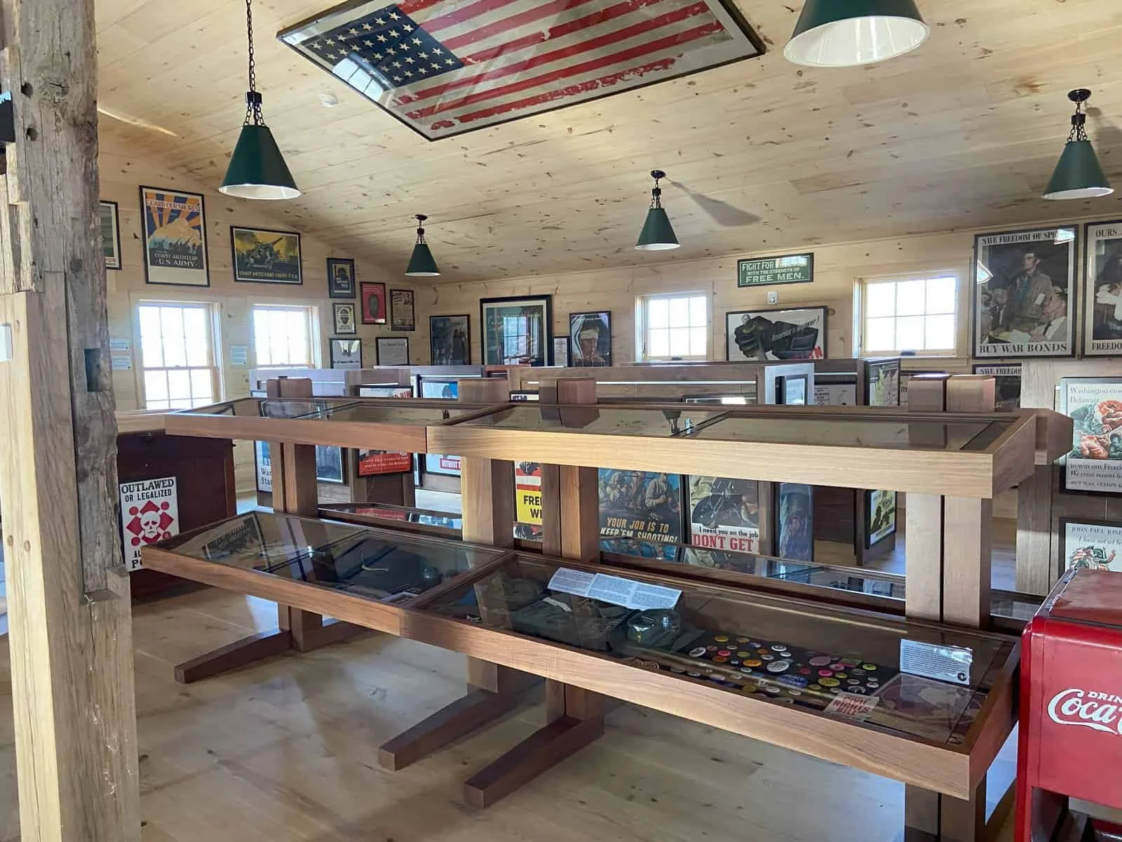 Museum interior with display cases of artifacts and vintage posters on wooden walls, American flag artwork on ceiling, and hanging green pendant lights.