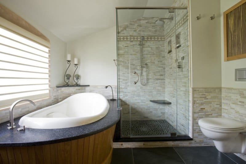 Bathroom with a large white bathtub, a glass-enclosed shower with a rain showerhead, and a toilet. The room has light-colored tiled walls and a window with horizontal blinds.