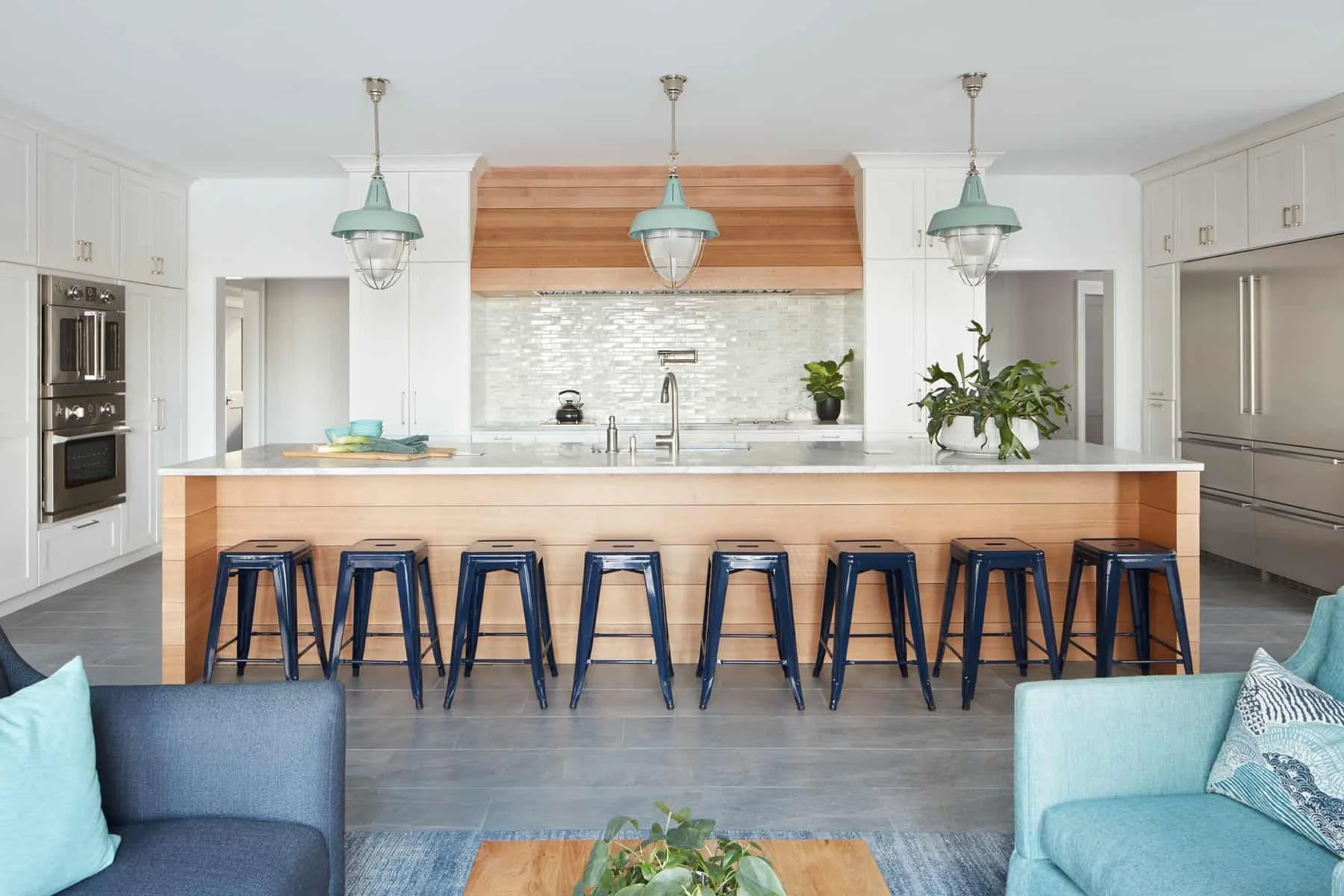 Modern kitchen with a large island, six blue bar stools, white cabinetry, stainless steel appliances, a stone backsplash, and decorative plants. There are three pendant lights hanging above the island.
