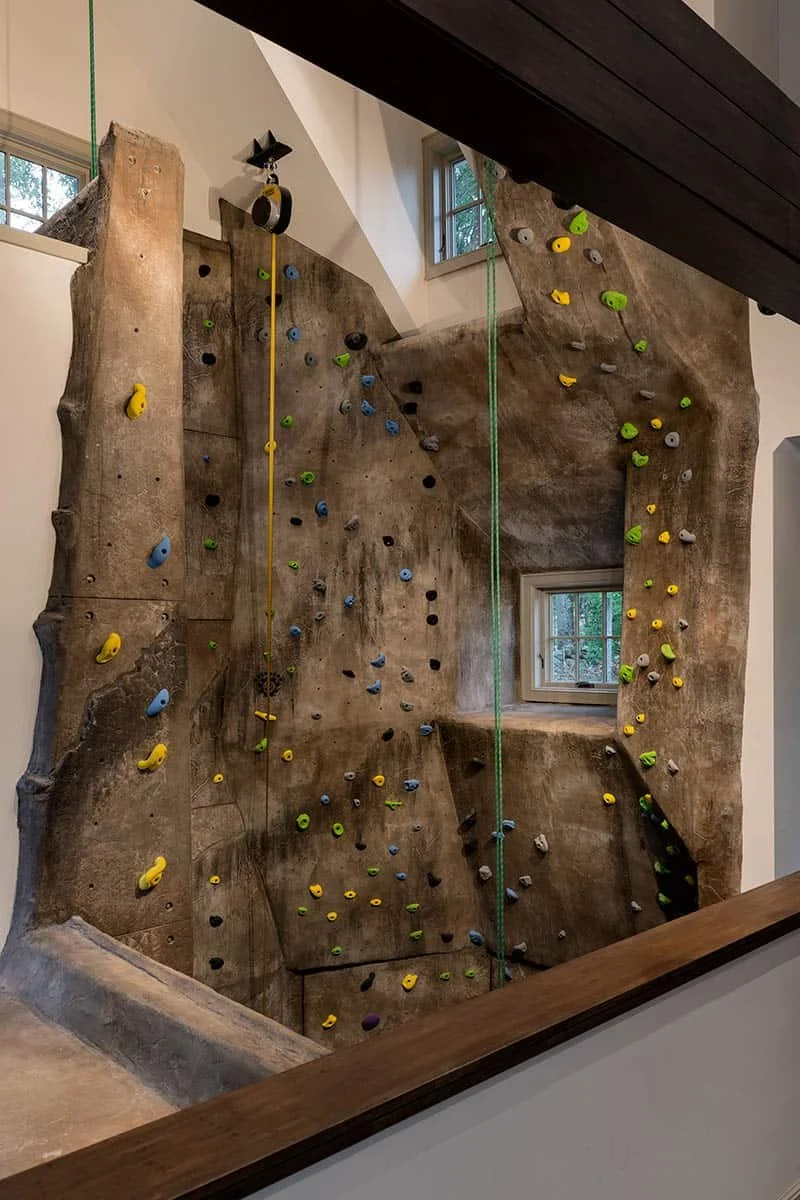 Indoor rock climbing wall with various colored holds and two windows, viewed from above a wooden railing.