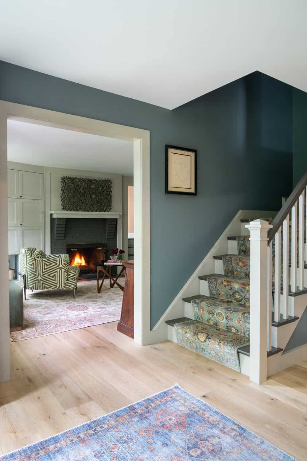 Interior view of a home's entryway leading to a living room with a fireplace and patterned armchair, featuring a staircase with a floral-patterned carpet runner.