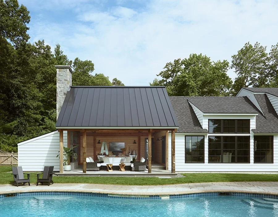 Backyard of a house with a pool, outdoor seating, and a covered patio area with wooden support beams, surrounded by trees.