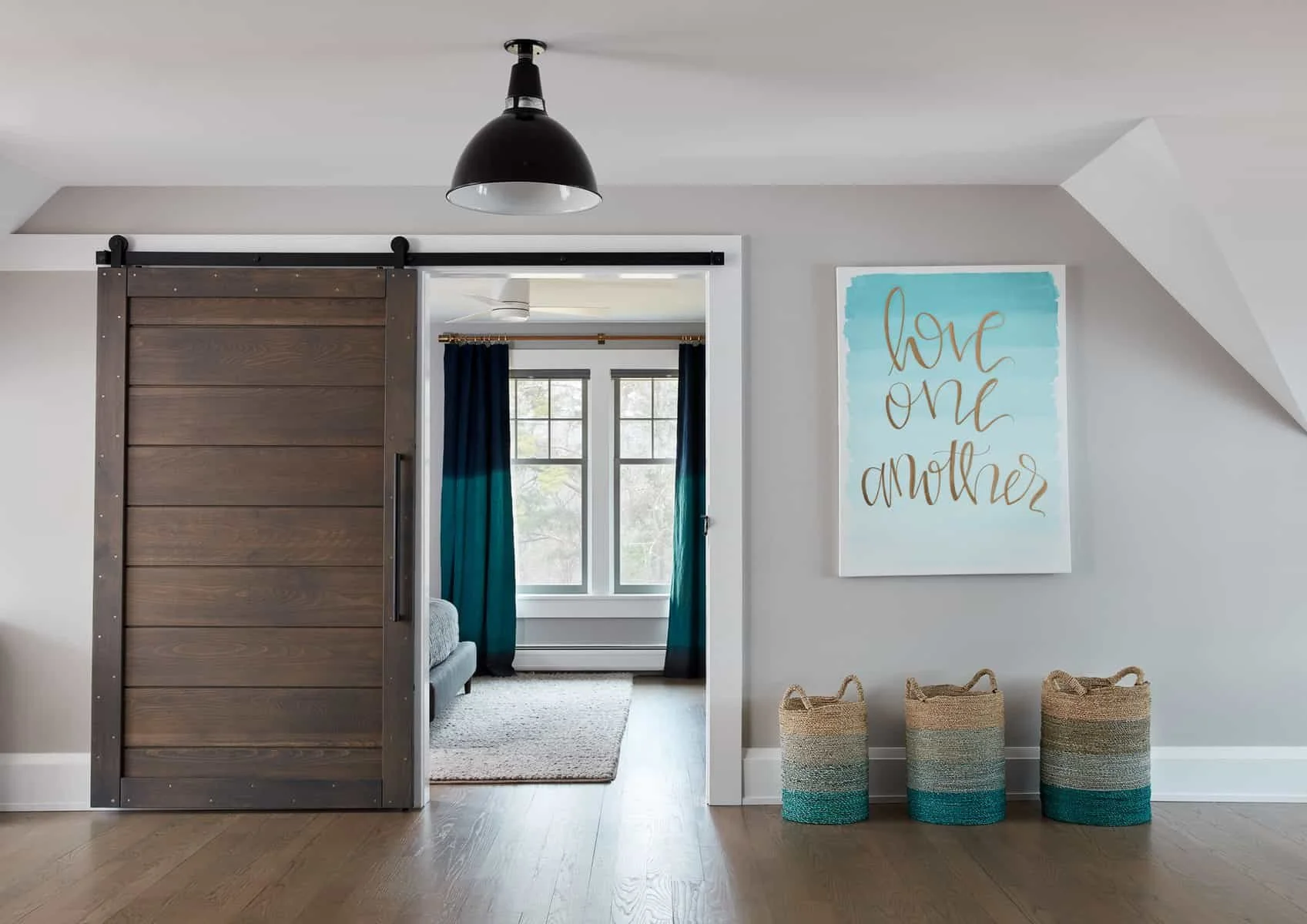 Interior of a room with a sliding barn door, a large window with dark curtains, a wall sign that says 'love one another,' and three woven storage baskets on the floor.