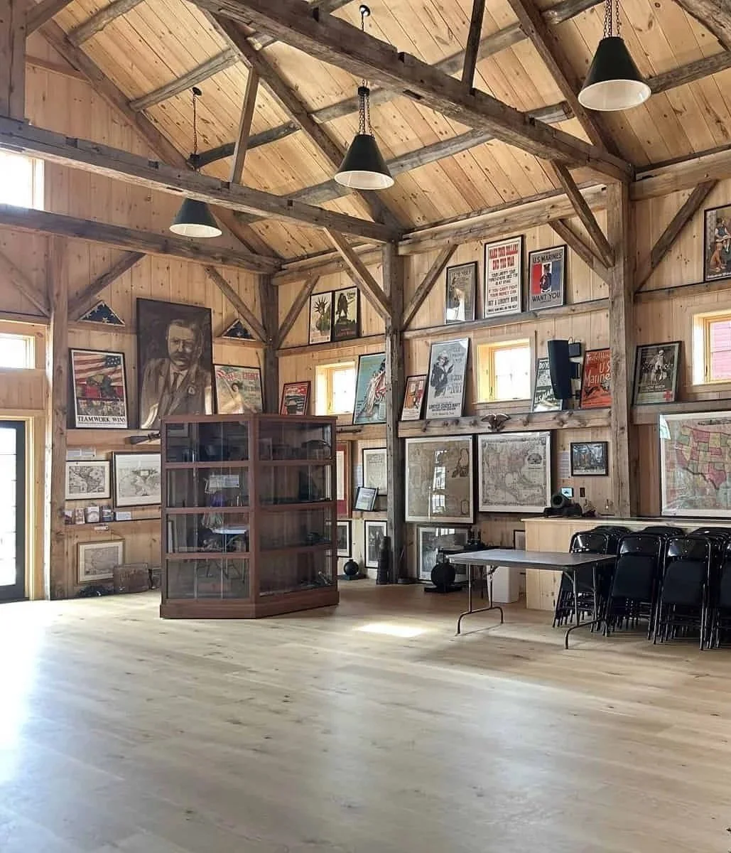A large, rustic wooden room with framed posters and pictures on the walls, wooden beams on the ceiling, and a display cabinet in the center. There are several chairs stacked against the right wall and a small table near the display cabinet.