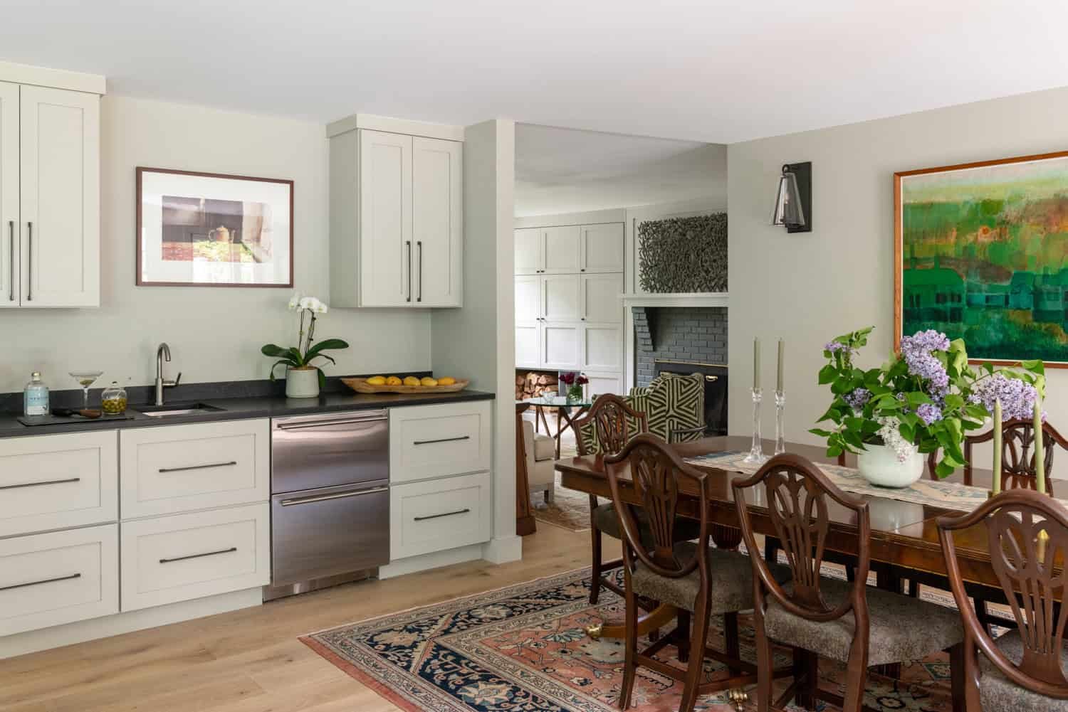 A bright, modern kitchen with white cabinets, black countertops, and a stainless steel dishwasher. There is a dining table with eight wooden chairs, a vase with purple flowers, and a framed colorful painting on the wall. The space includes a cozy sit