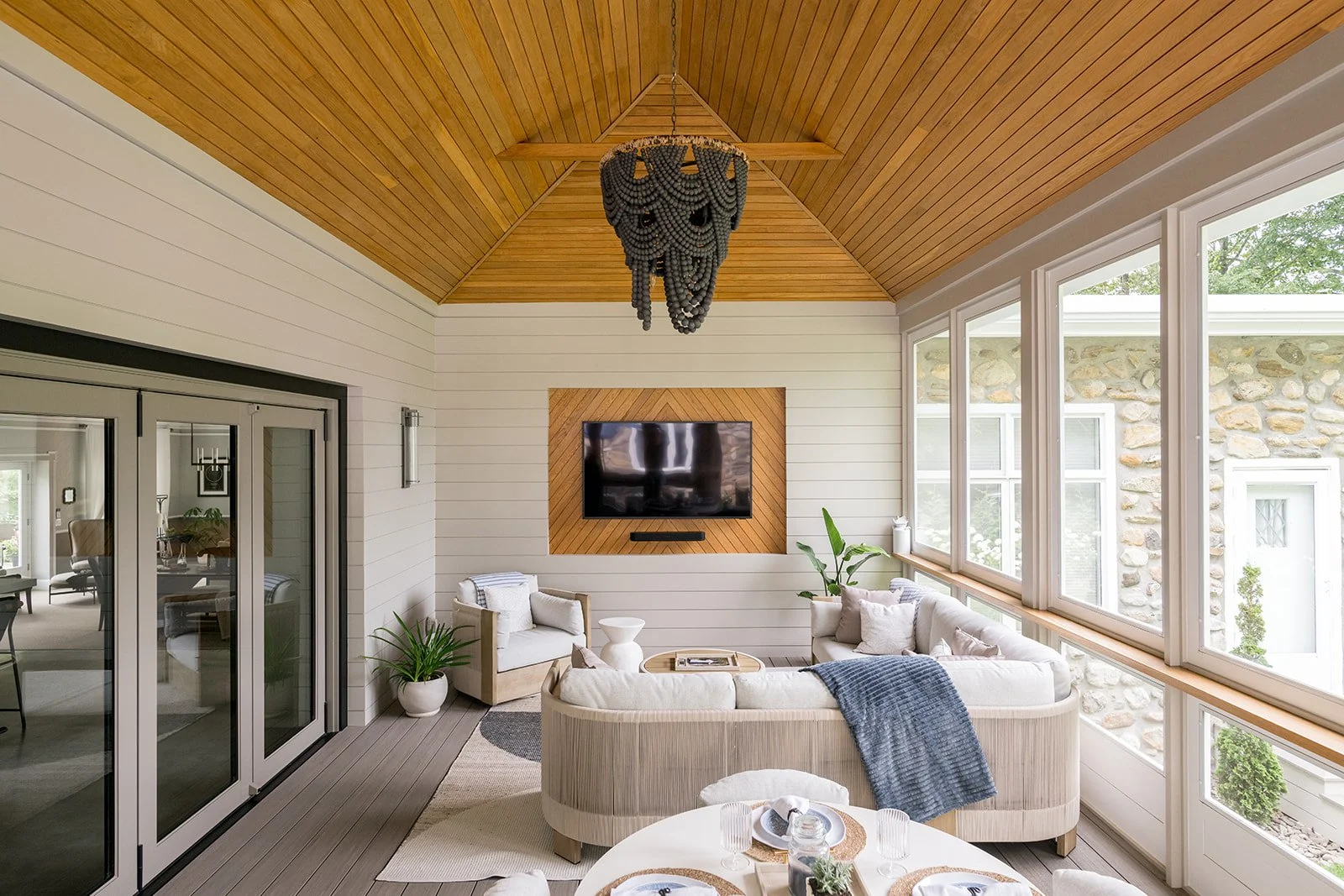 Sunroom with wood ceiling, white walls, large windows, modern chandelier, wall-mounted TV, white sofa with cushions, potted plants, tied blanket, and sliding glass door leading to indoor space.