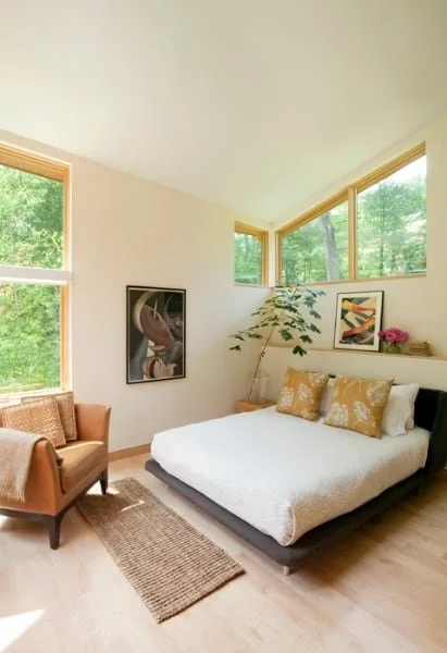 Bright bedroom with a bed, decorative pillows, a plant, framed art, a pink flower, and a cozy armchair near large windows with green trees outside.