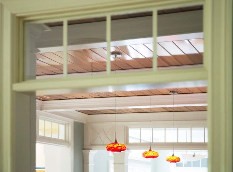 View through a window showing a room with a wooden ceiling, three hanging red and yellow pendant lights, and windows allowing natural light.