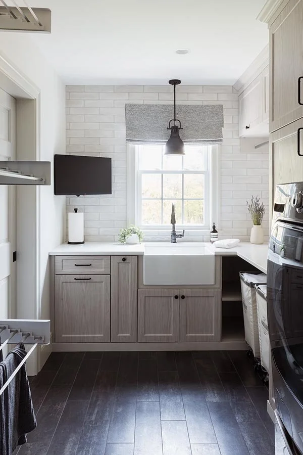 Bright laundry room with white brick wall, gray cabinets, laundry sink, and window with gray valance, black pendant light, and small plants.