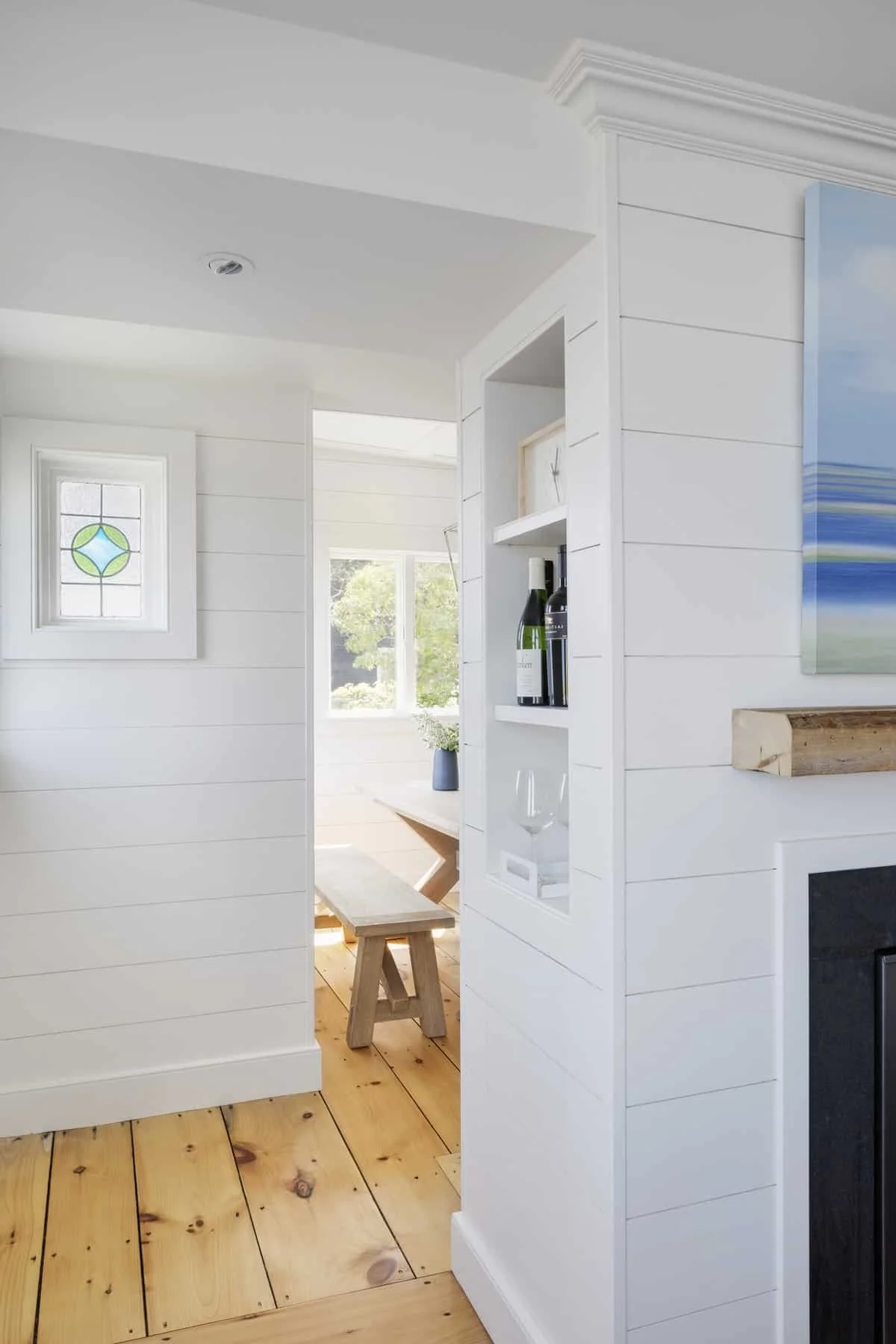 Interior view of a bright, modern living space with white shiplap walls, wooden flooring, a small window with stained glass, and a glimpse into a dining area with a table, bench, and potted plant, showcasing minimalist decor.
