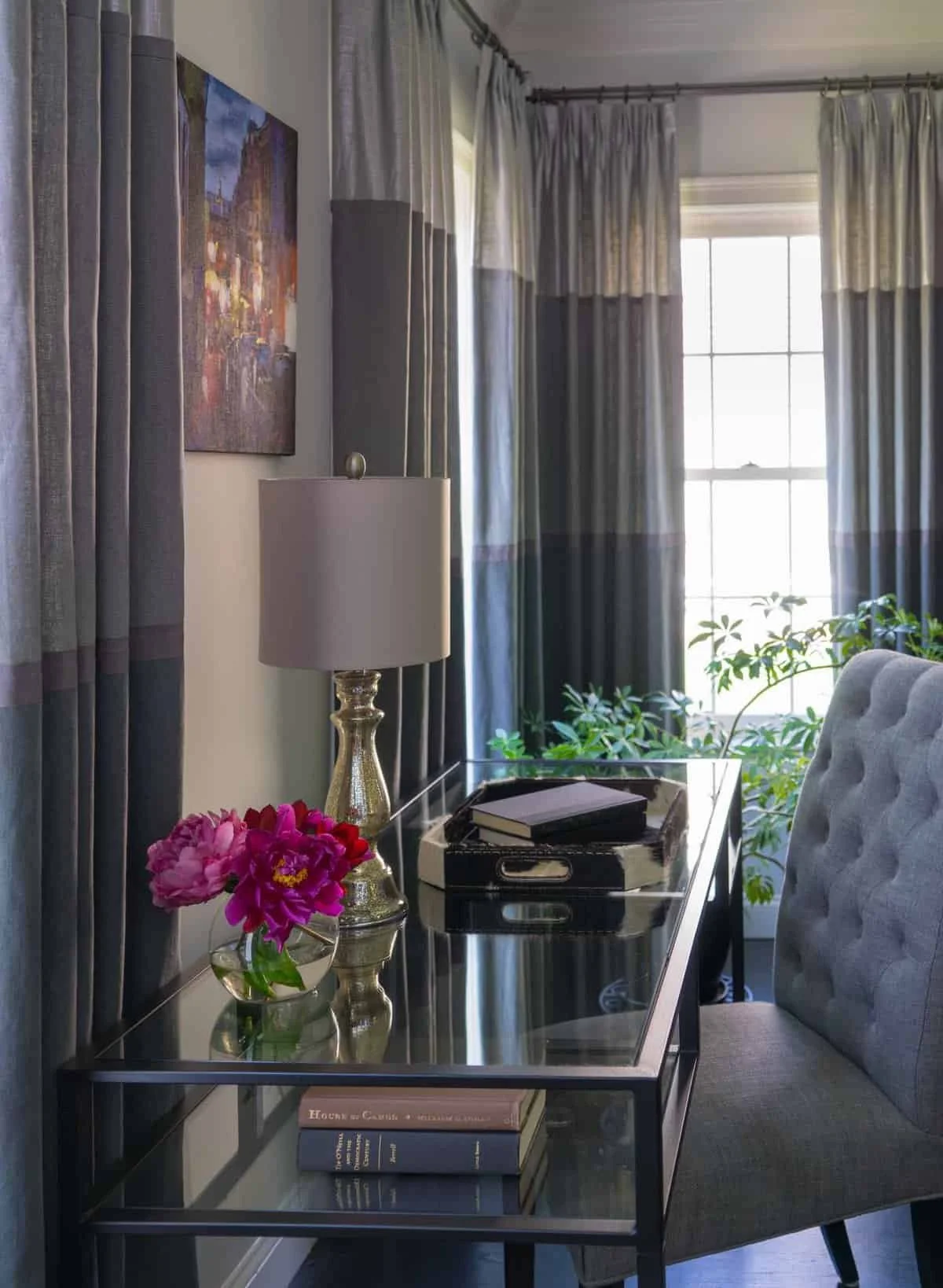 Interior living room corner with a glass-top side table, a beige upholstered armchair, a table lamp with a metallic base, a small vase with pink flowers, a tray with books, a window with light-colored curtains, and a painting on the wall. Green plant
