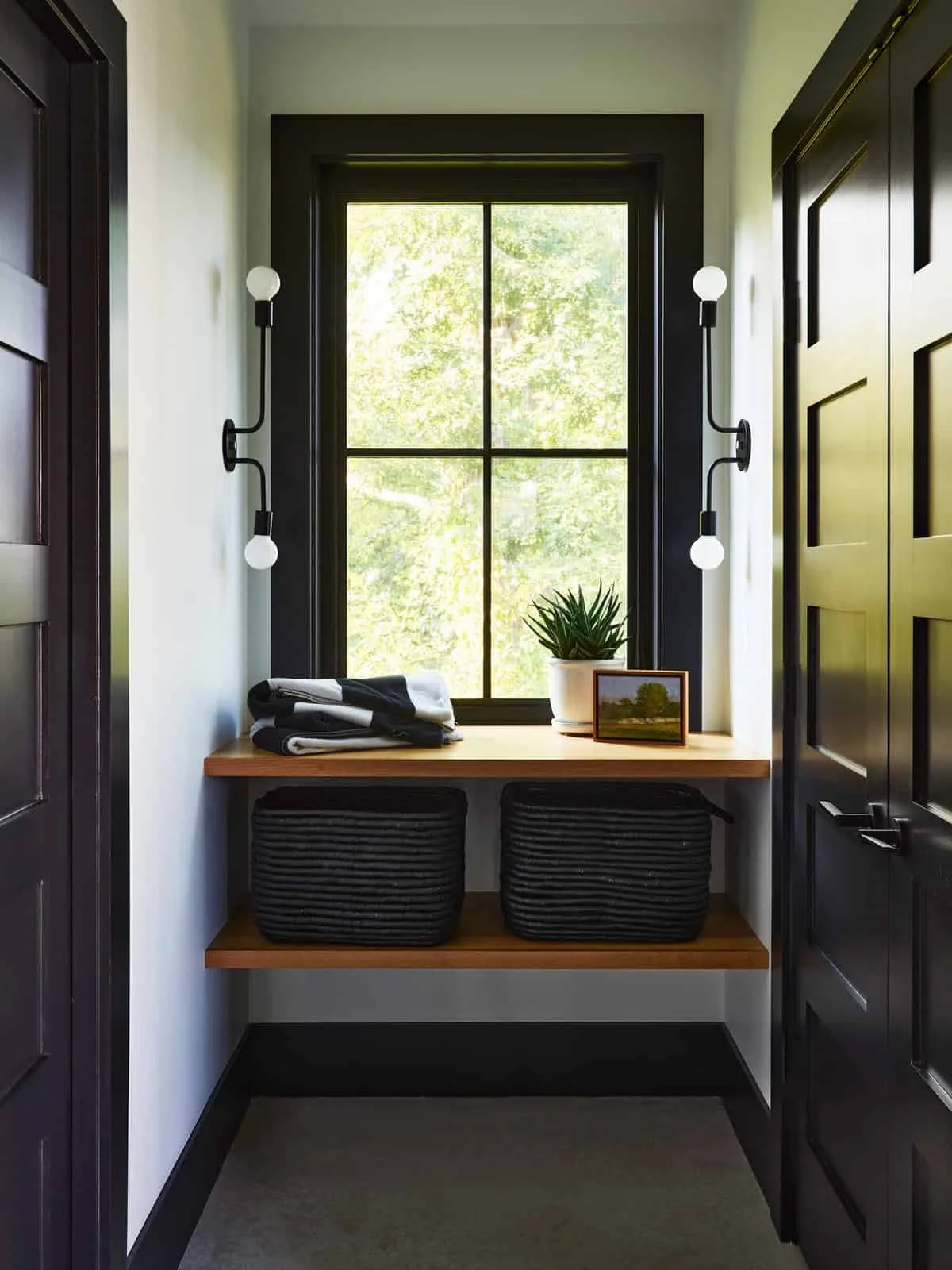 Corner of a room with a window, a wooden shelf below the window with folded black and white towels, a potted plant, and a small framed picture. Two black wall-mounted light fixtures with round bulbs are on each side of the window.
