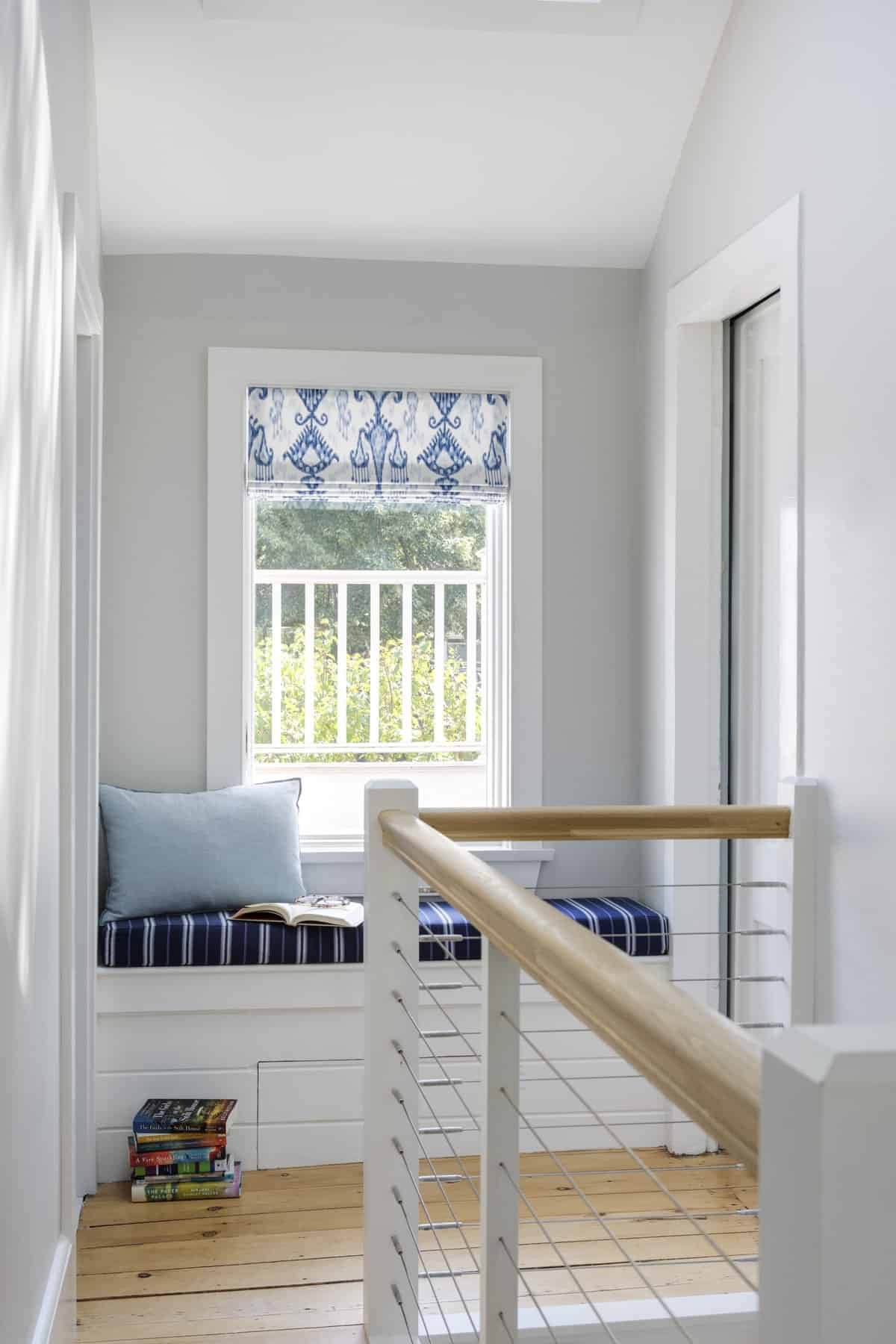A cozy nook by a window with a blue and white striped cushion, a light blue pillow, an open book, and a small stack of books underneath. The window has a blue patterned curtain, and outside the window, there is a view of green trees and a white raili