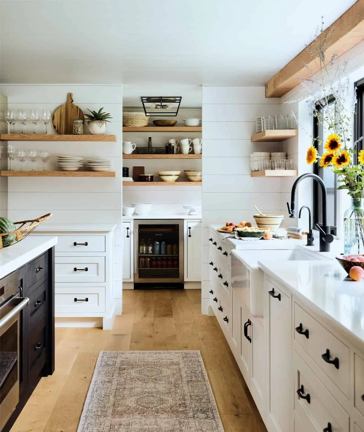 A bright farmhouse-style kitchen with white cabinets, wooden open shelves holding dishes and glassware, a black faucet at the sink with sunflowers in a vase, and light hardwood flooring.