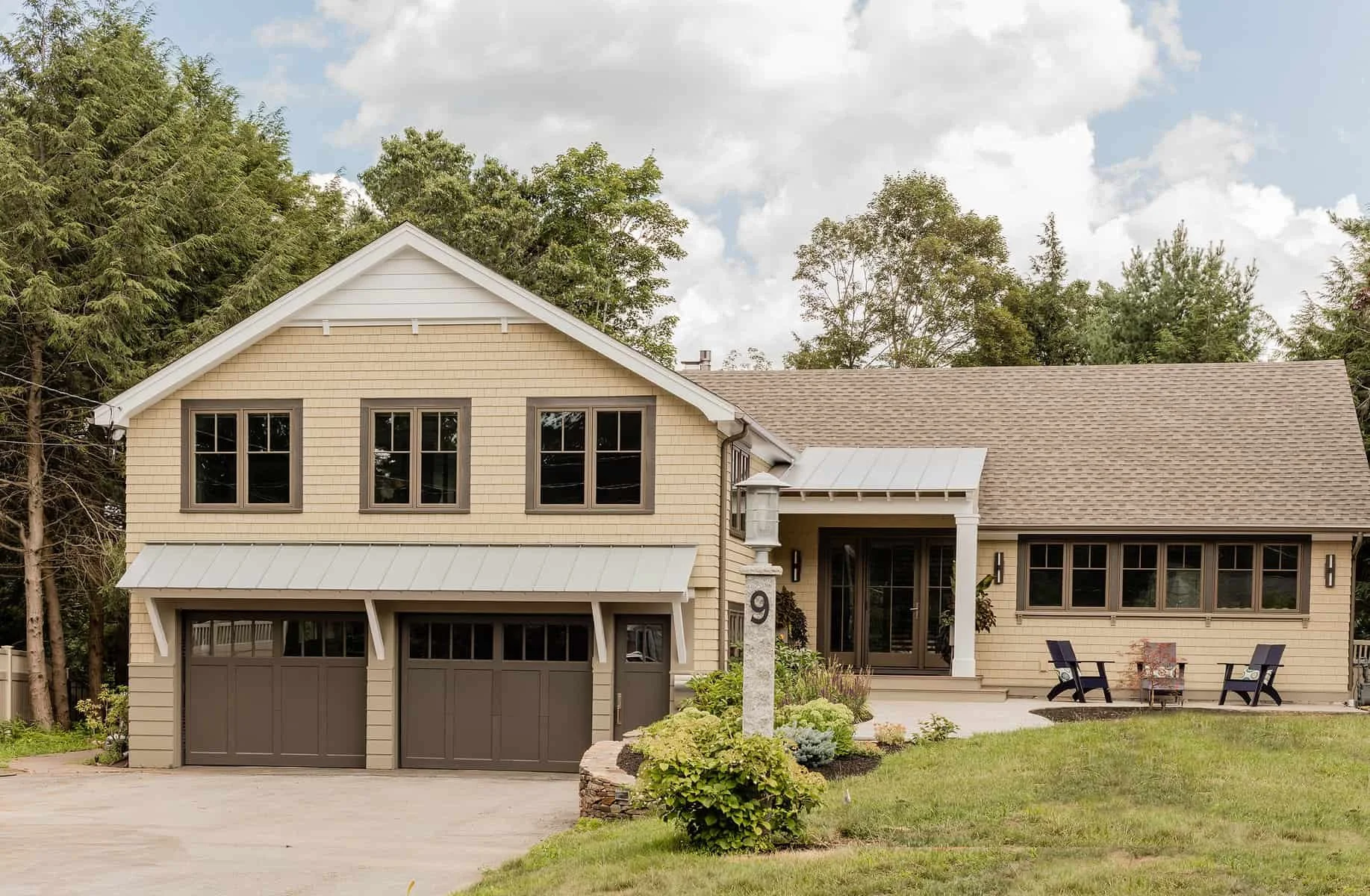Front view of a two-story house with a beige exterior, brown garage doors, and a covered porch with outdoor seating. The house has multiple windows, a sloped roof, and is surrounded by green lawn and trees.