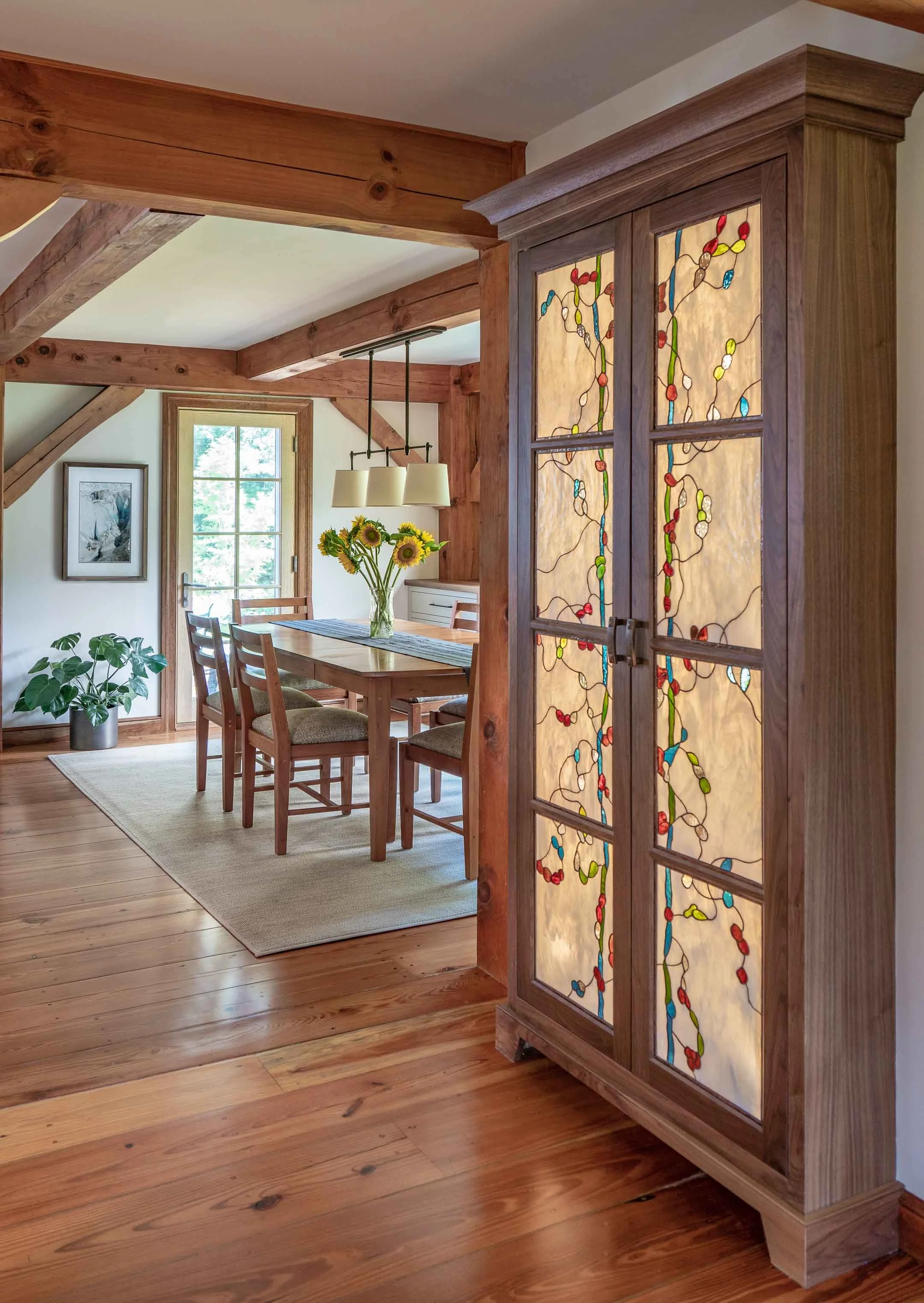 A wooden room features a stained glass cabinet with colorful abstract patterns, a dining table with chairs, a window, a hanging light fixture, and a vase with sunflowers on the table.