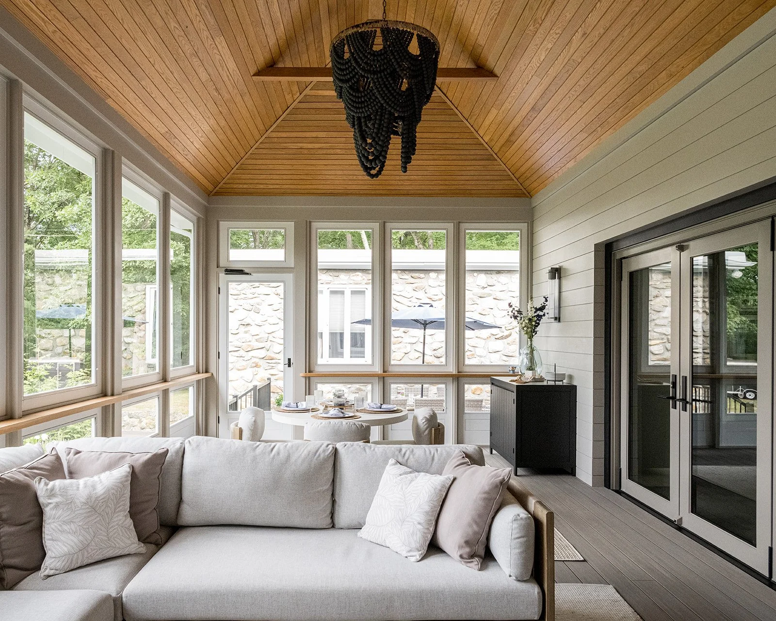 Living room with large windows, a white sectional sofa with pillows, a round dining table with chairs, a black cabinet with a vase of flowers, and a black chandelier hanging from the wooden vaulted ceiling.