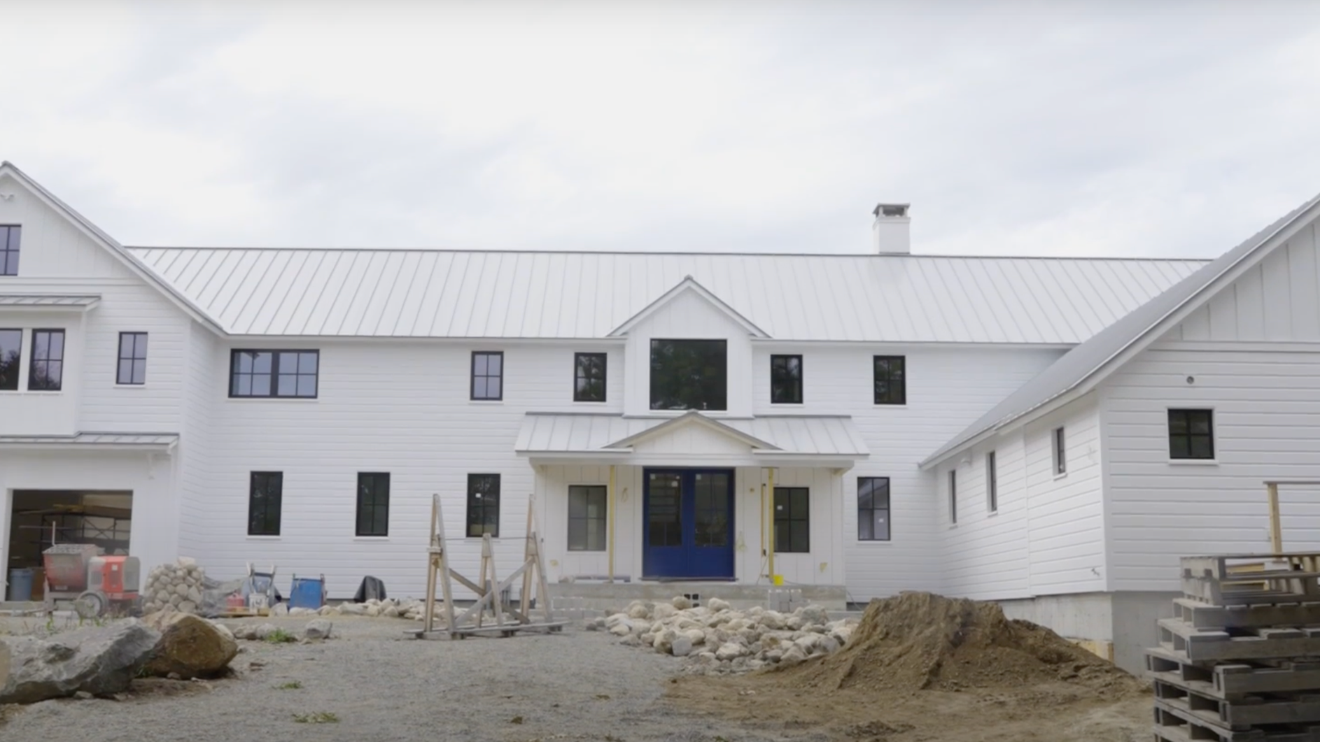 Under construction white multi-story custom modern farmhouse with blue front door, surrounded by construction materials and dirt, with cloudy sky overhead.