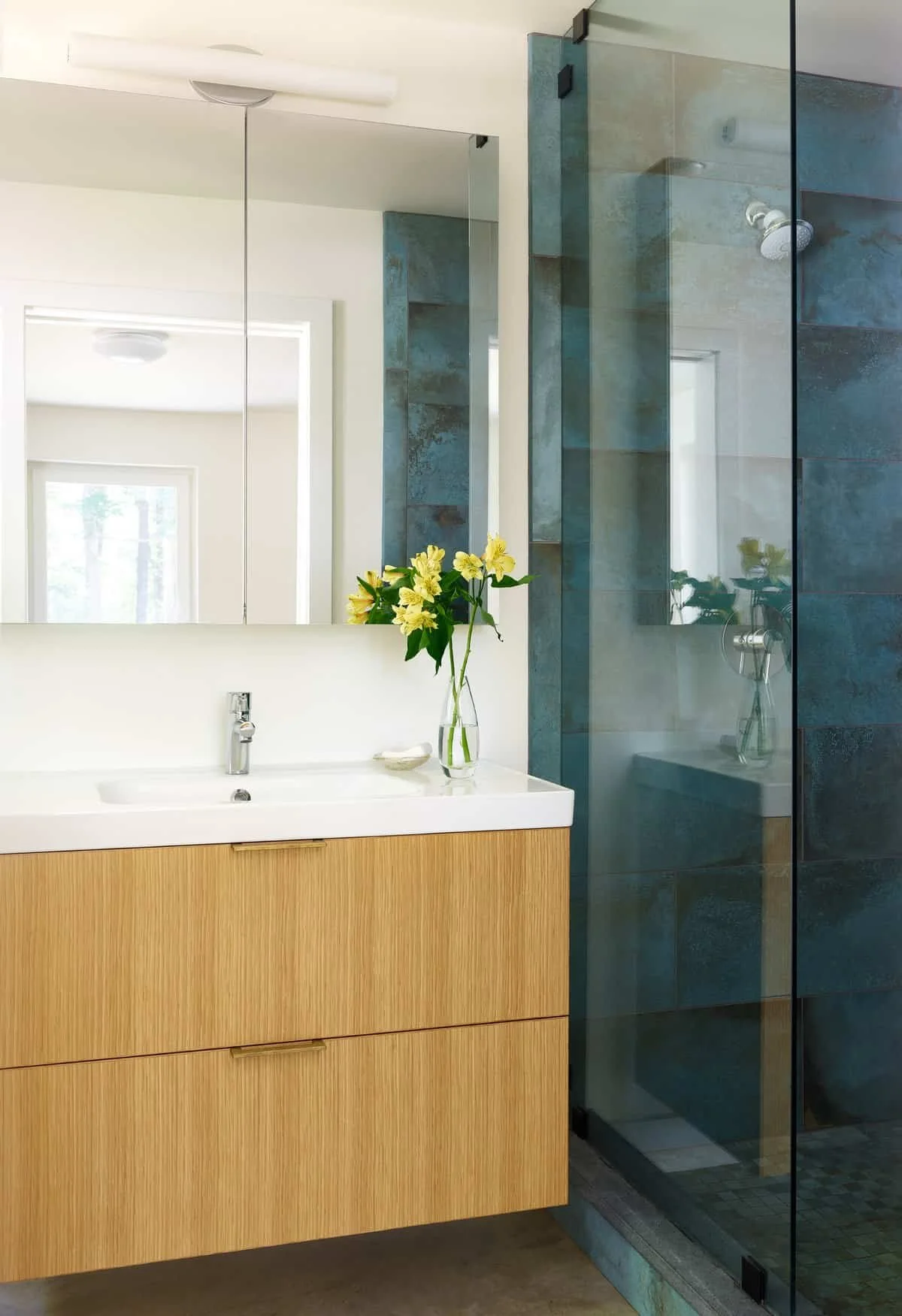 A modern bathroom features a white countertop with a wooden vanity, a sink with a silver faucet, and a glass vase with yellow flowers. A large mirror reflects part of the room, and a separate glass-enclosed shower with dark tiles and a showerhead is 