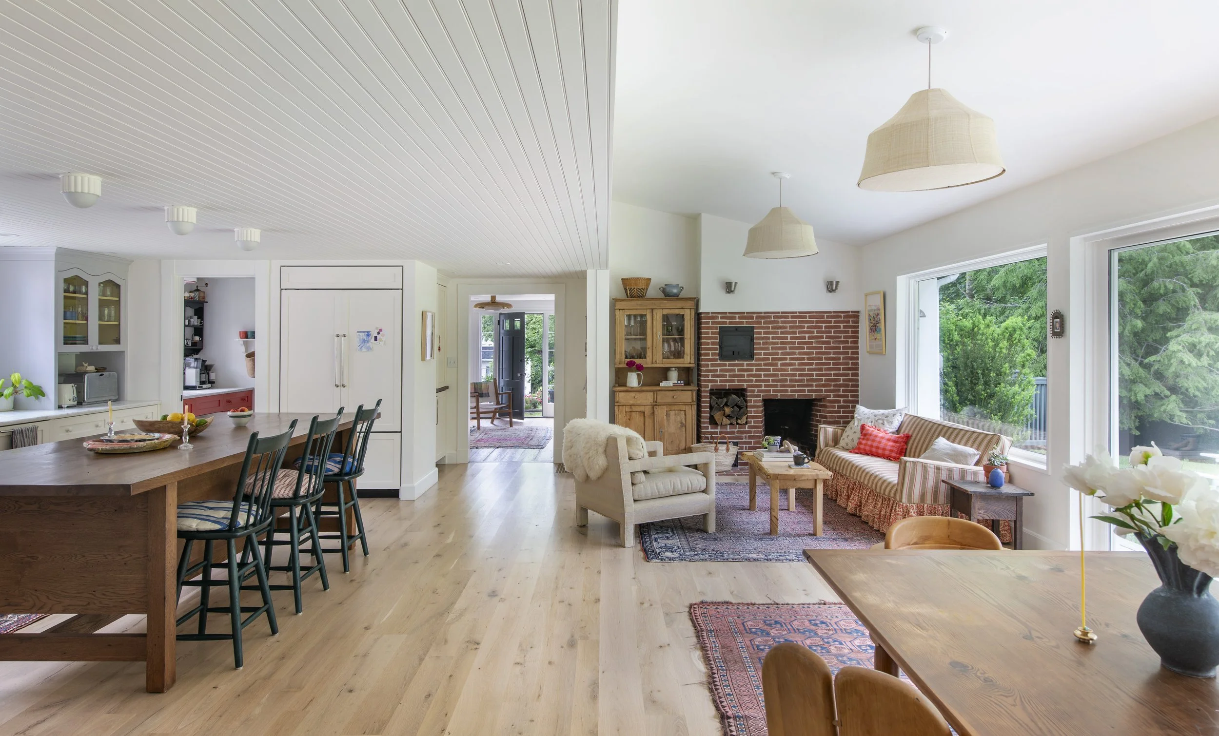 Bright, open living and dining room with white walls, hardwood floors, large windows, a brick fireplace, cozy seating, and a dining table with chairs in a stylish, modern decor.
