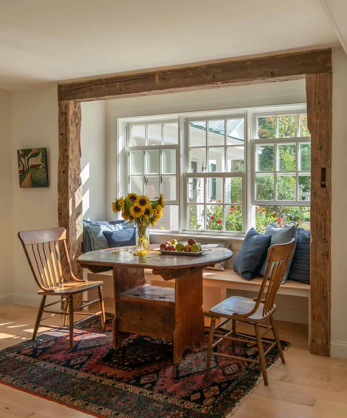 Sunlit dining nook with a wooden table, sunflower centerpiece, and cushioned window seat with pillows, surrounded by rustic wood beams and a patterned area rug.