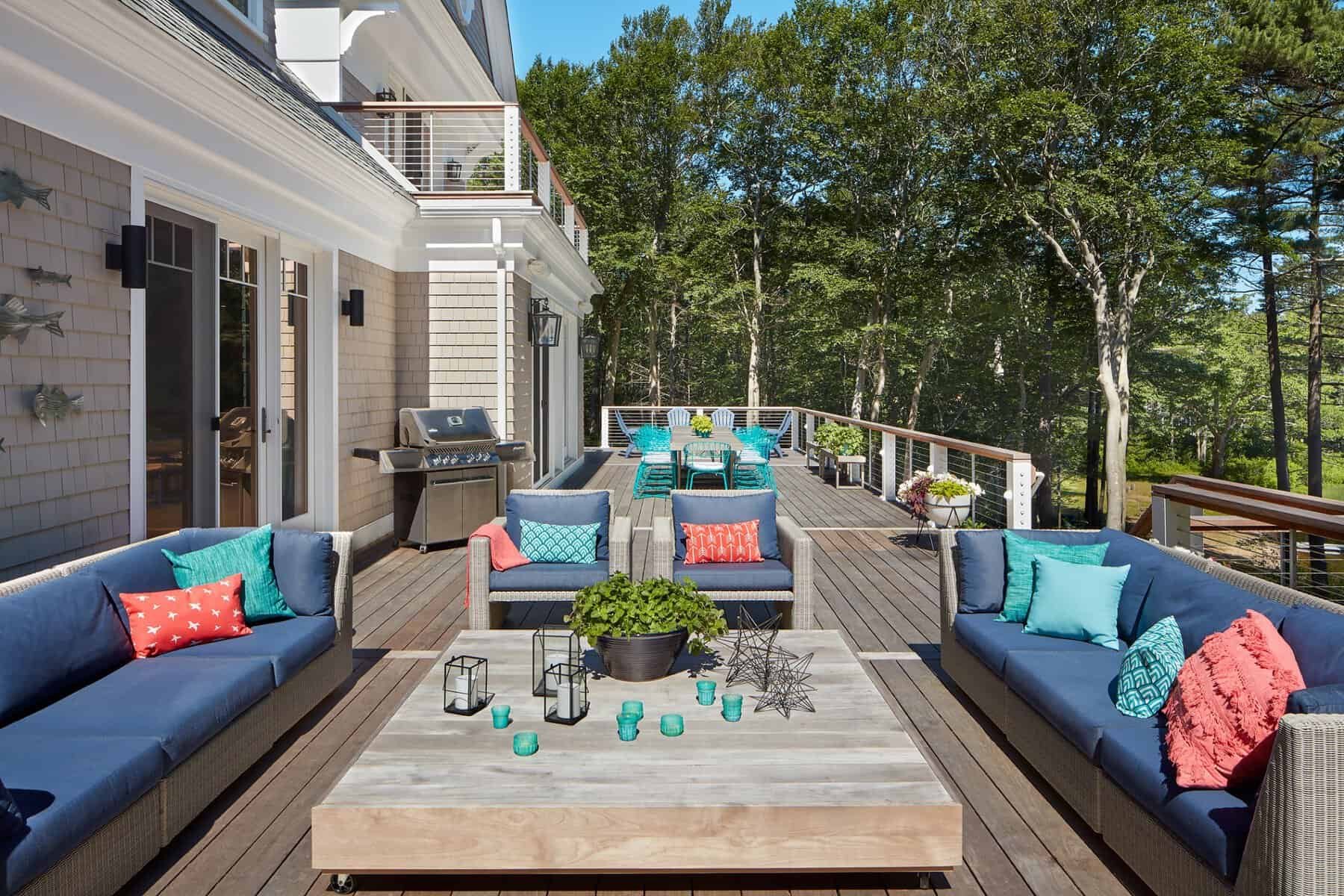 A spacious outdoor deck with navy blue sofas decorated with colorful cushions, a large glass-top coffee table with candles and decorative items, a dining area with teal chairs and a table, a grill, and a scenic view of tall trees in the background.
