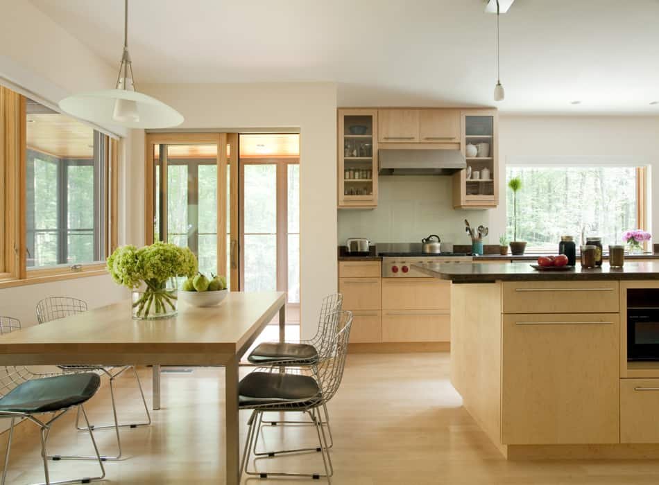Kitchen with wooden cabinets, a central island, and stainless steel appliances. A dining table with chairs and a vase of green flowers is in the foreground, and large windows and sliding doors let in natural light.
